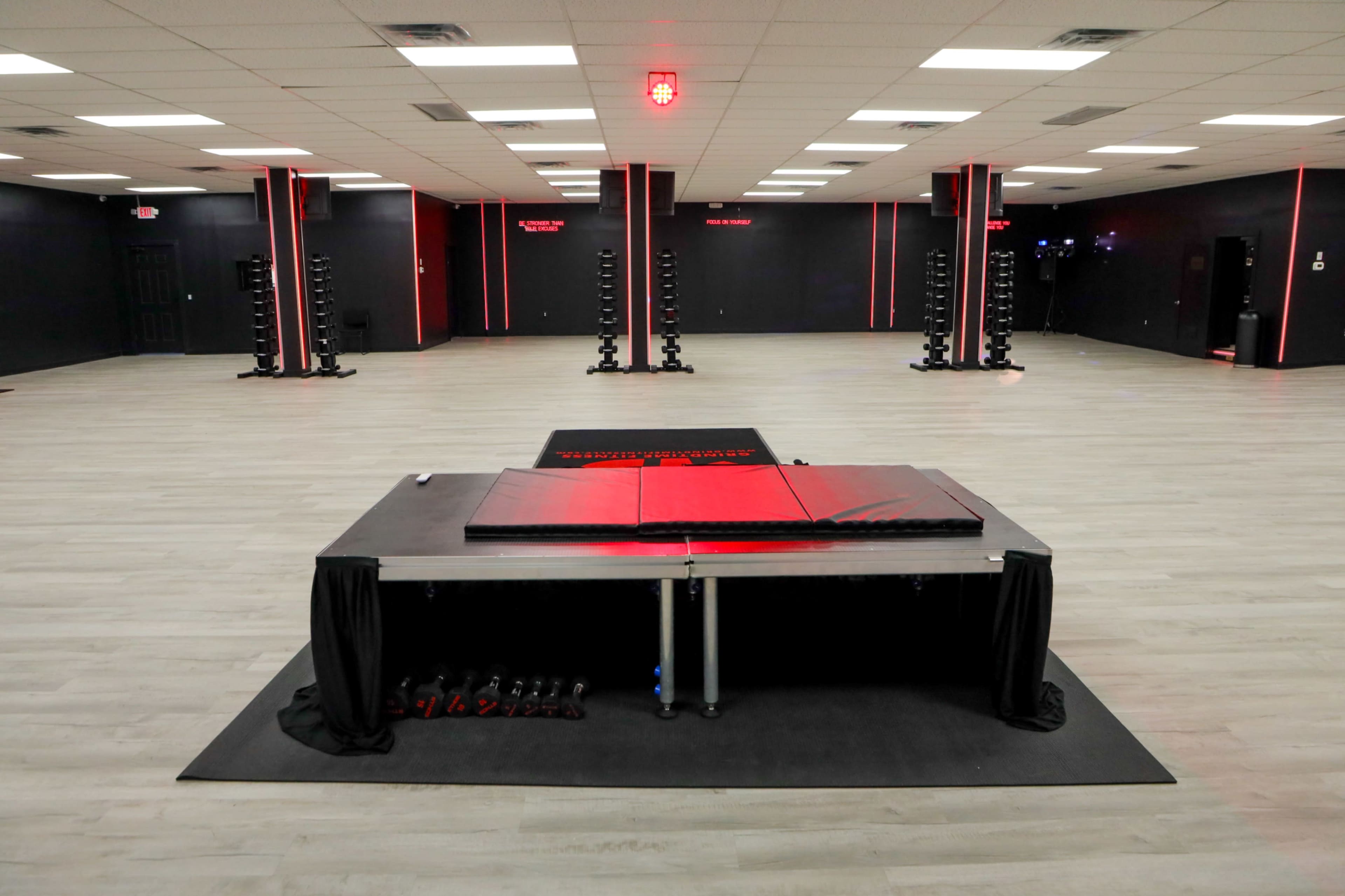 The image shows a large, empty gymnasium with gray flooring, red and black decor, and various exercise equipment arranged neatly along the walls.
