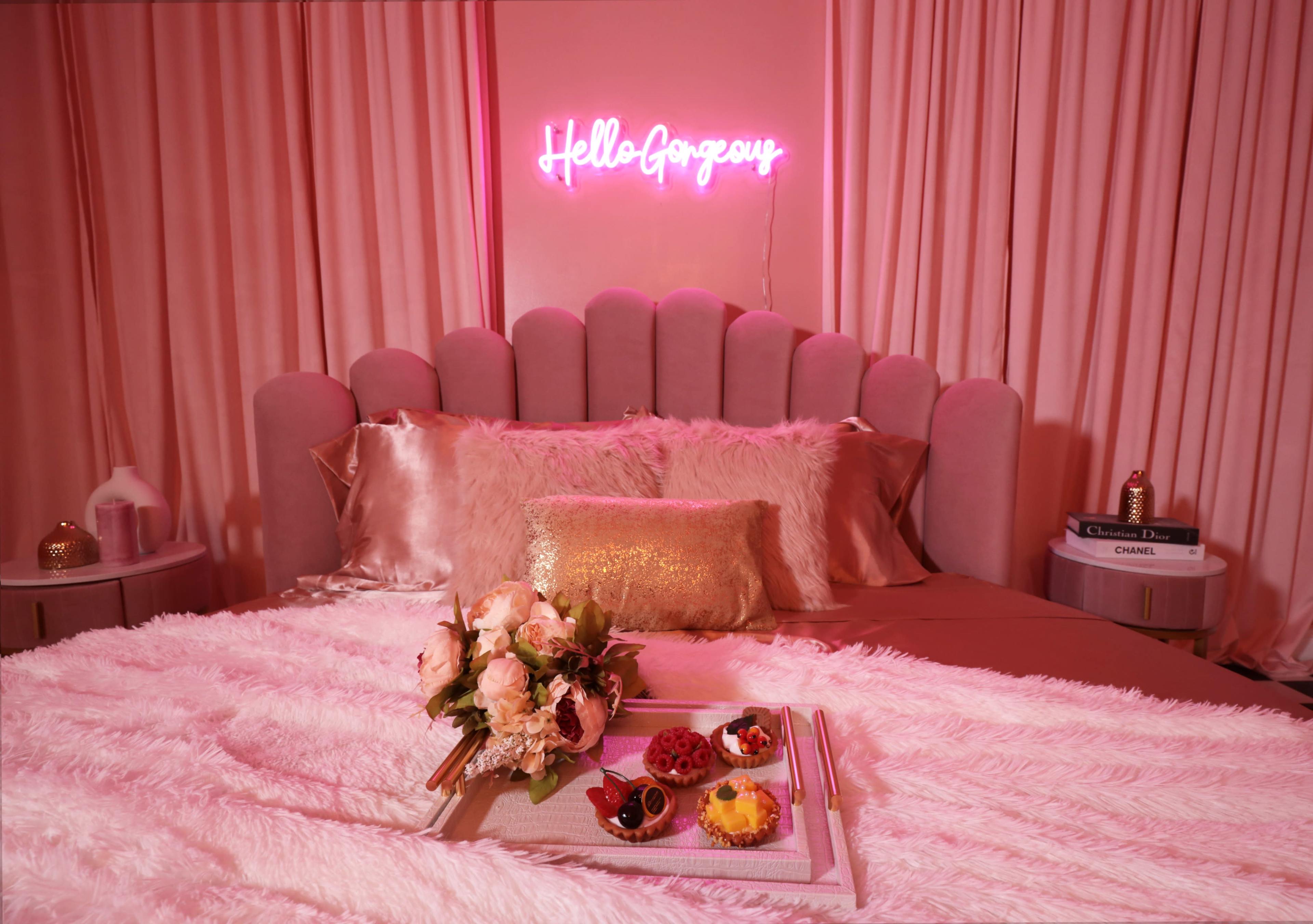 A pink-themed bedroom features a plush bed with a decorative headboard, a neon sign that says "Hello Gorgeous," and a tray with desserts and a bouquet of flowers on the bed.