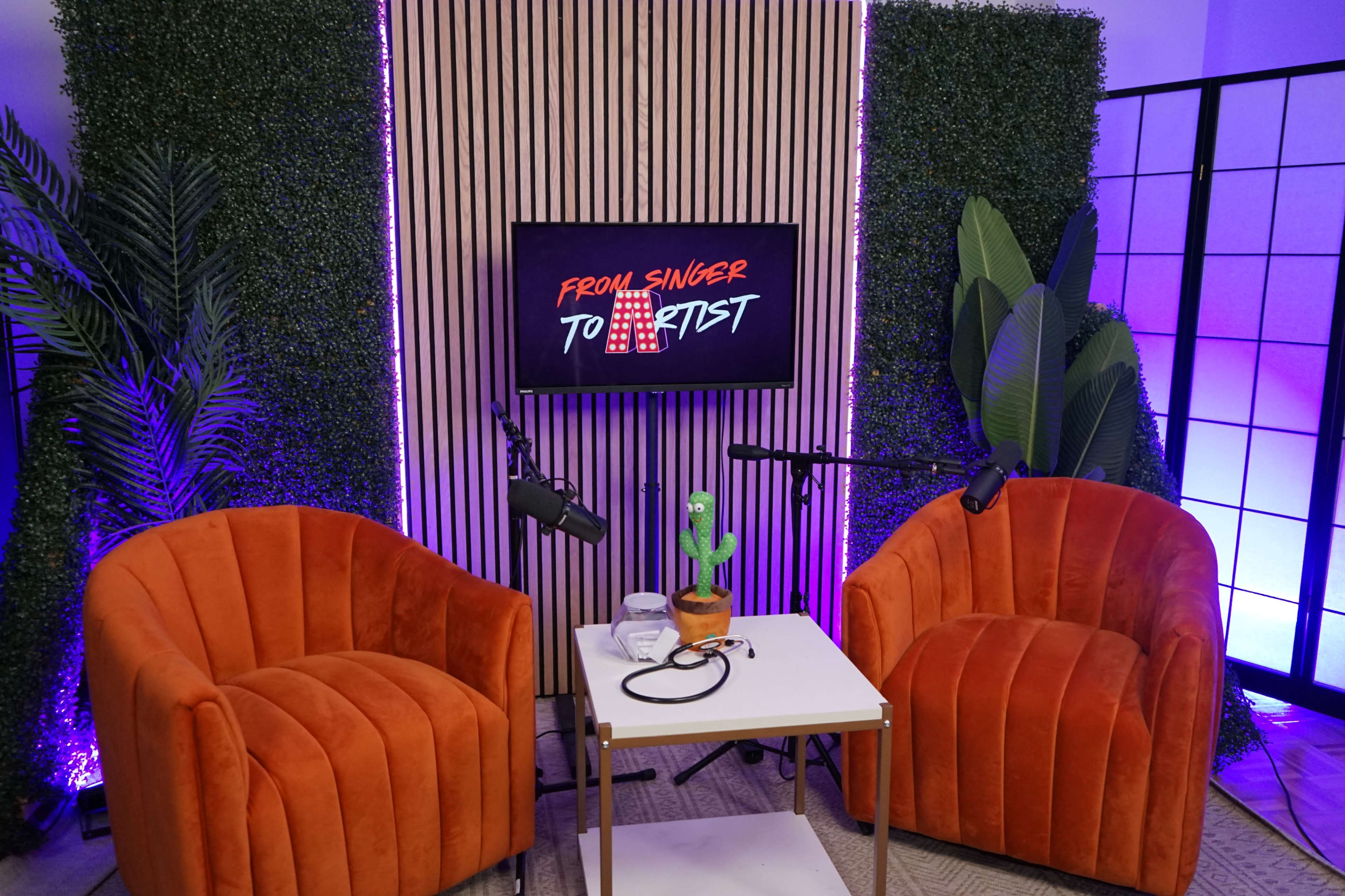 A set featuring two orange chairs, a small table with a plant and microphone cables, and a backdrop displaying the phrase "From Singer to Artist."