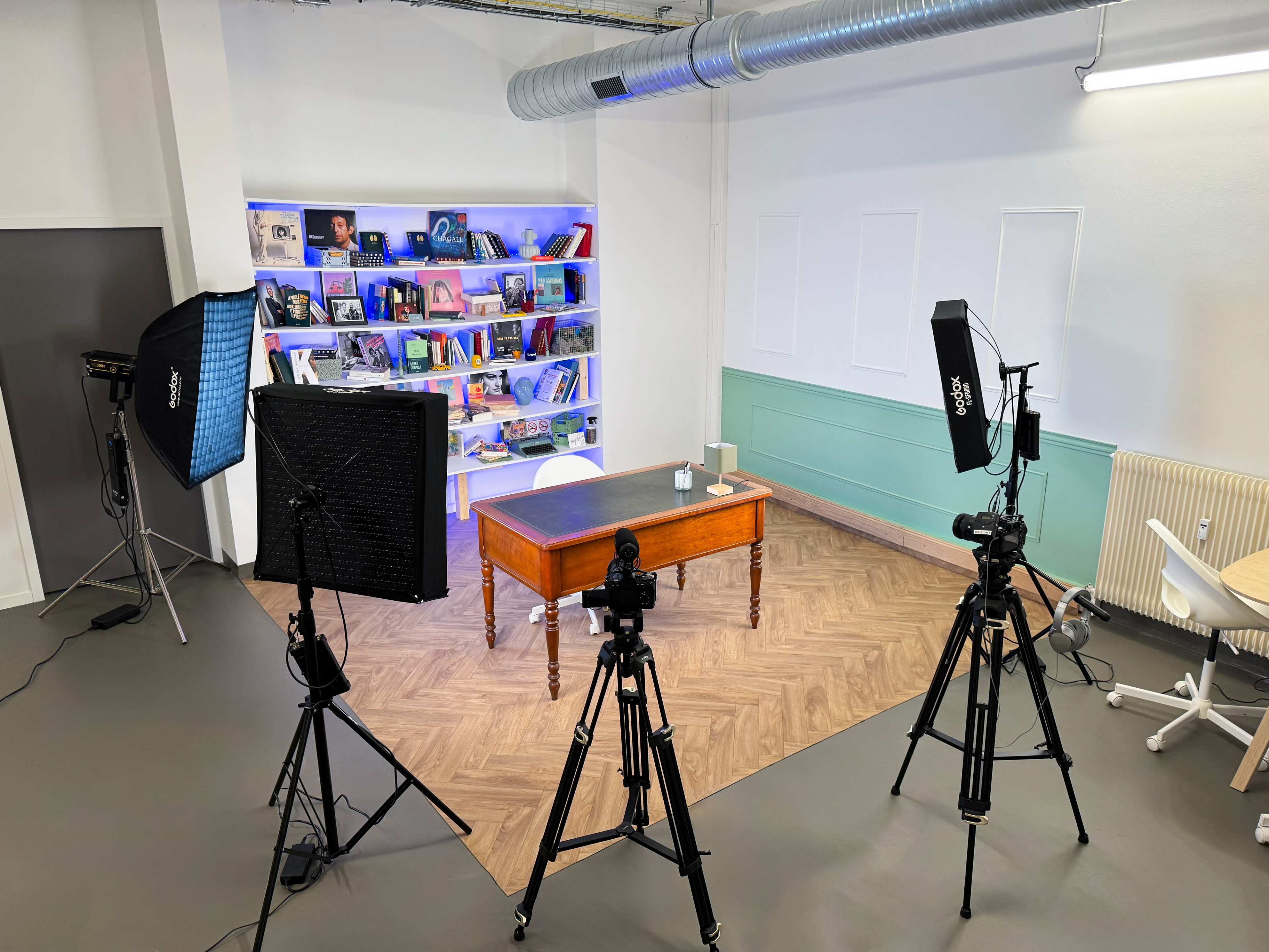 A well-lit studio setup features three tripods with cameras and softbox lights surrounding a wooden table, set against a background of shelves filled with books and decorative items.