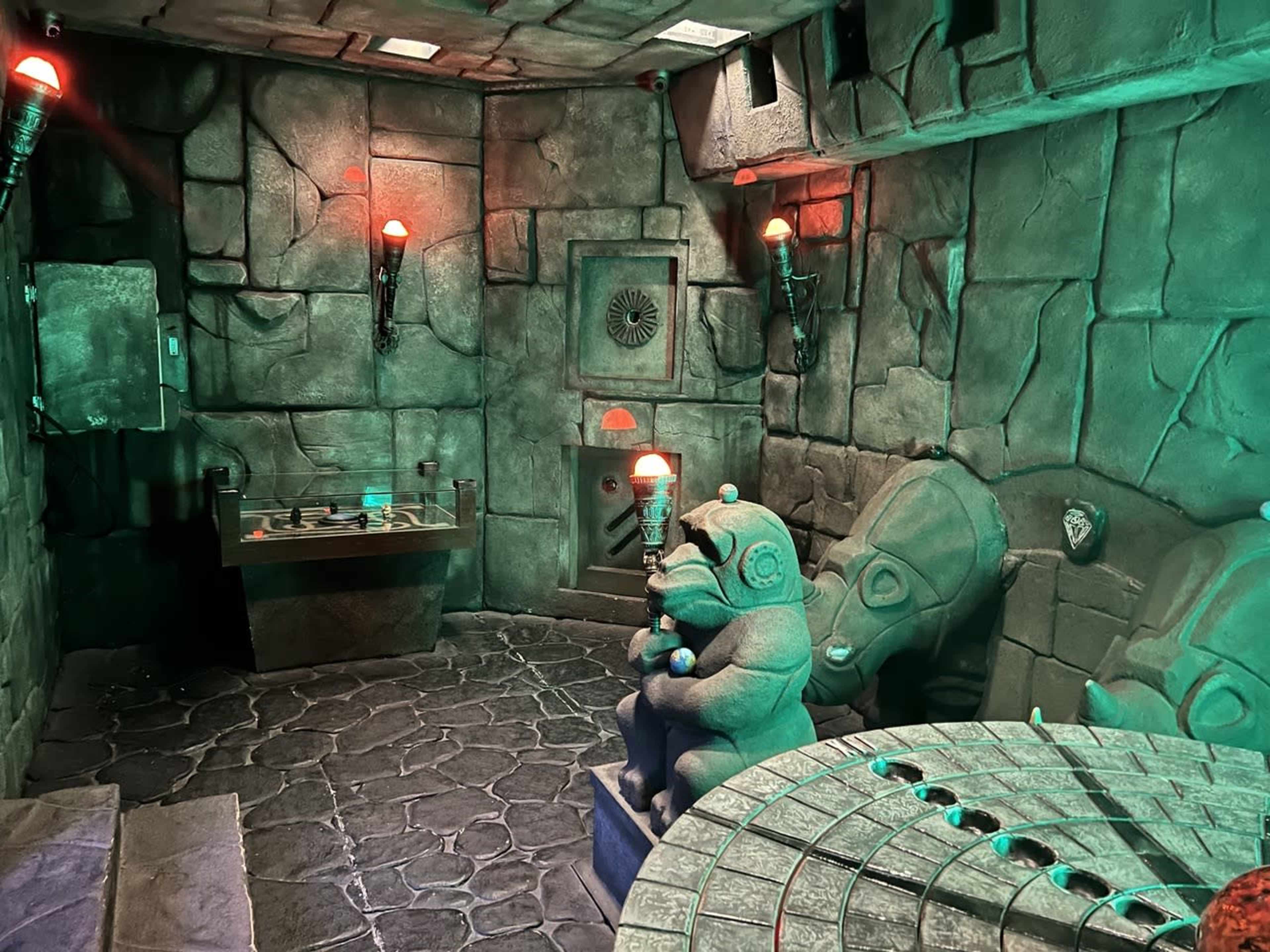 The image shows a dark, stone-walled room featuring ancient-looking sculptures, a circular stone table, and dimly lit lamps.