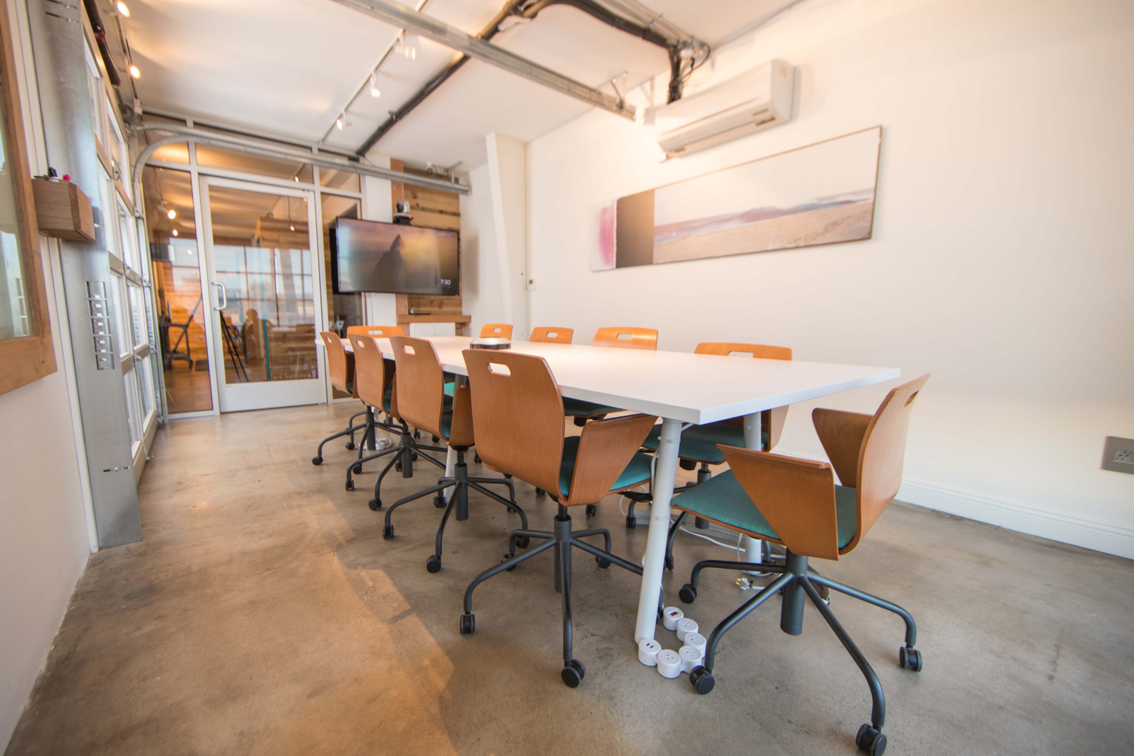 A modern conference room features a long white table surrounded by ergonomic wooden chairs and a large wall-mounted screen.