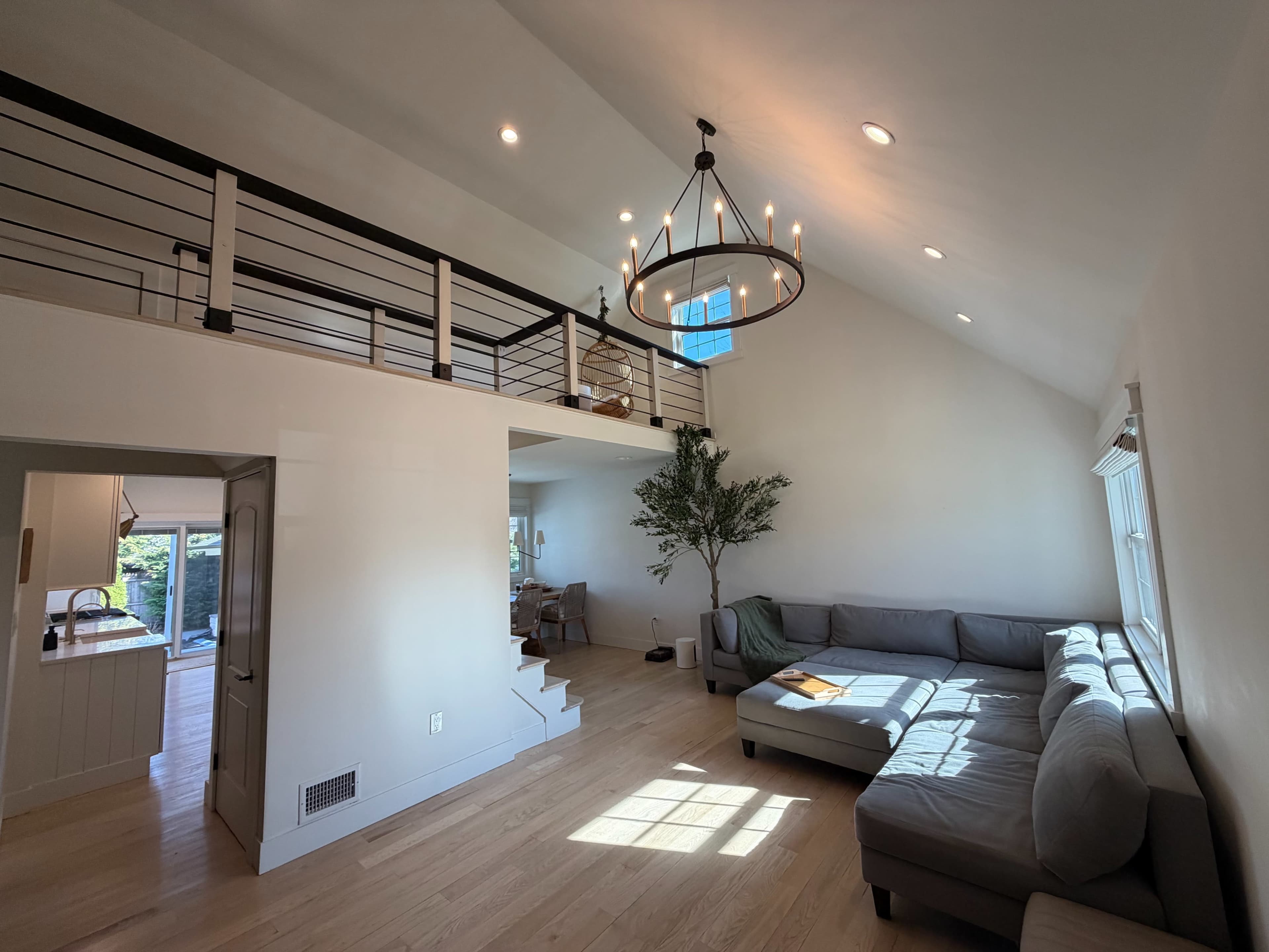 The image shows a bright, open living room with a large sectional couch, a tall chandelier, and a view of a dining area and kitchen beyond.