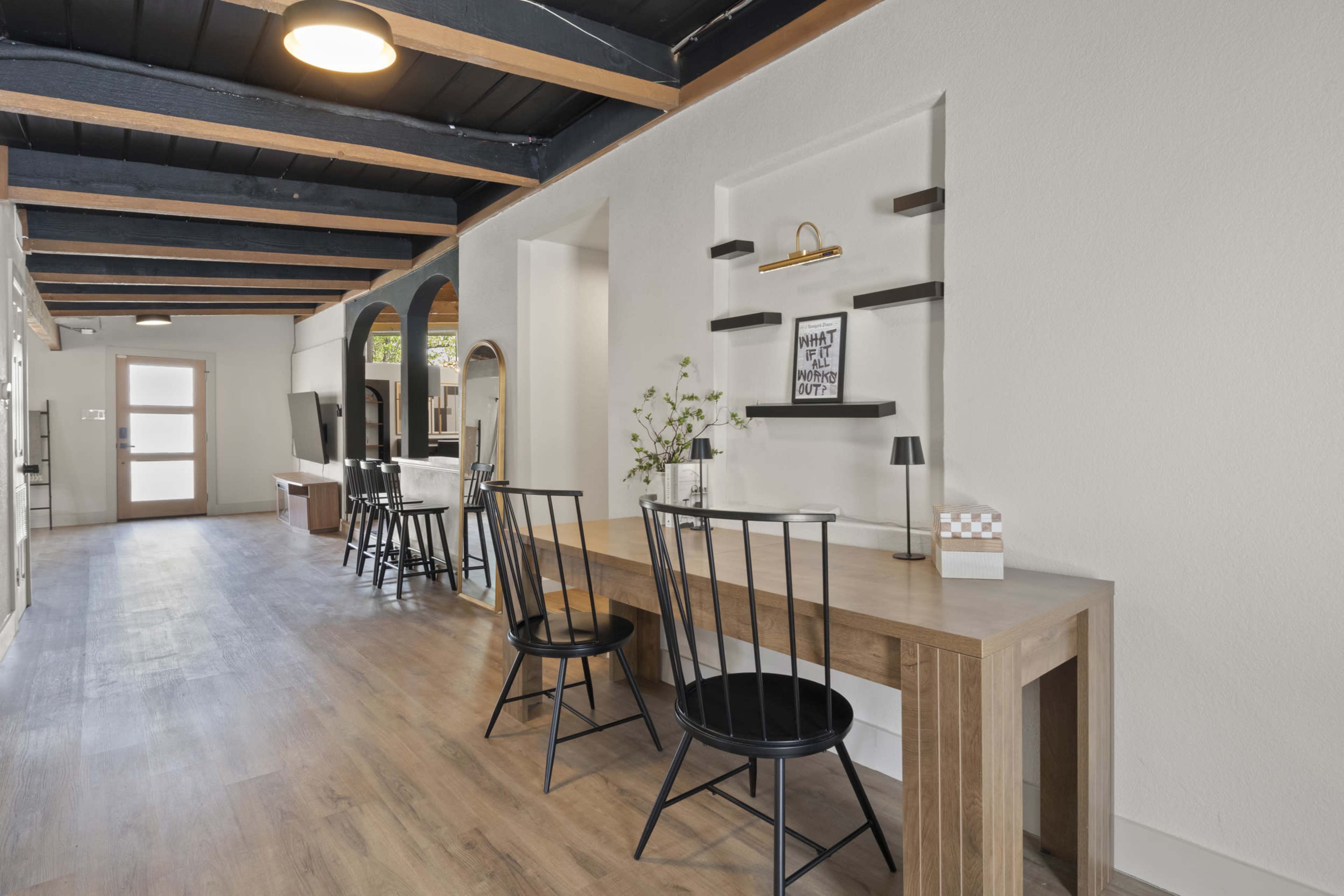 A long wooden table with black chairs is set against a wall in a spacious, modern interior with exposed beams and light flooring.