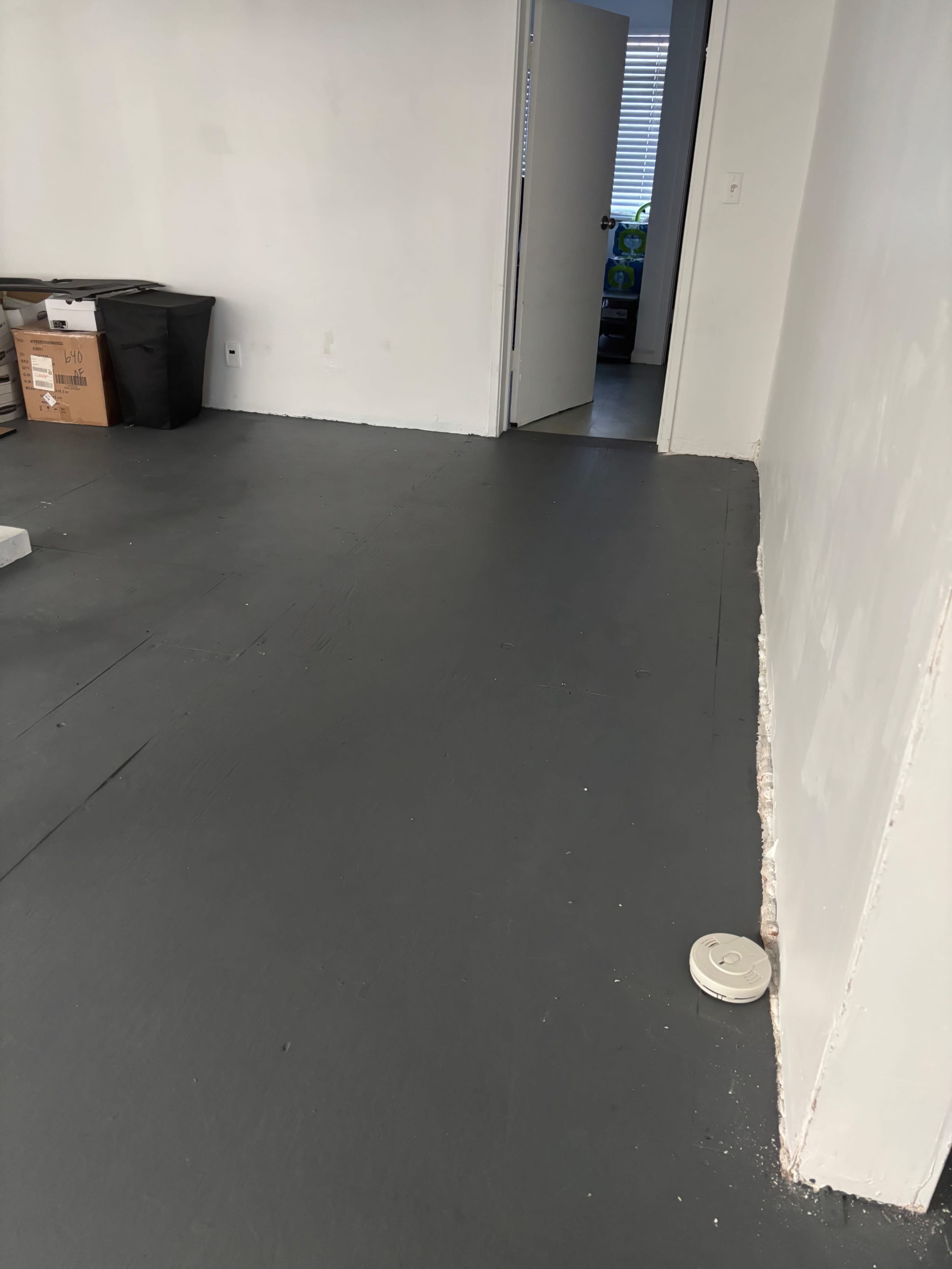 The image shows a sparsely furnished room with a gray floor, a door leading to another room, and a circular object on the floor near the wall.