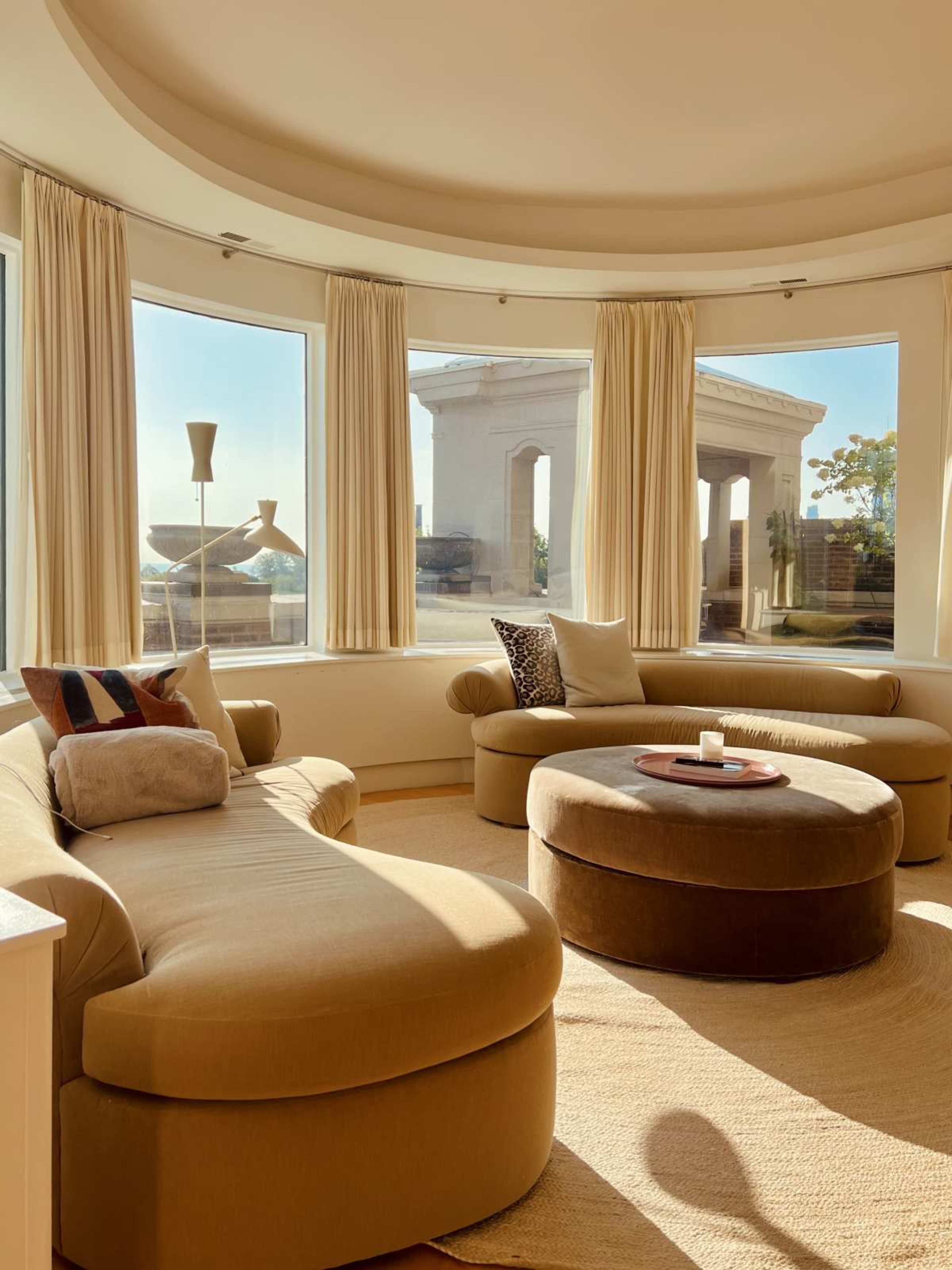 The scene features a circular, sunlit living room with two curved sofas and a round ottoman, facing large windows that showcase an outdoor view.