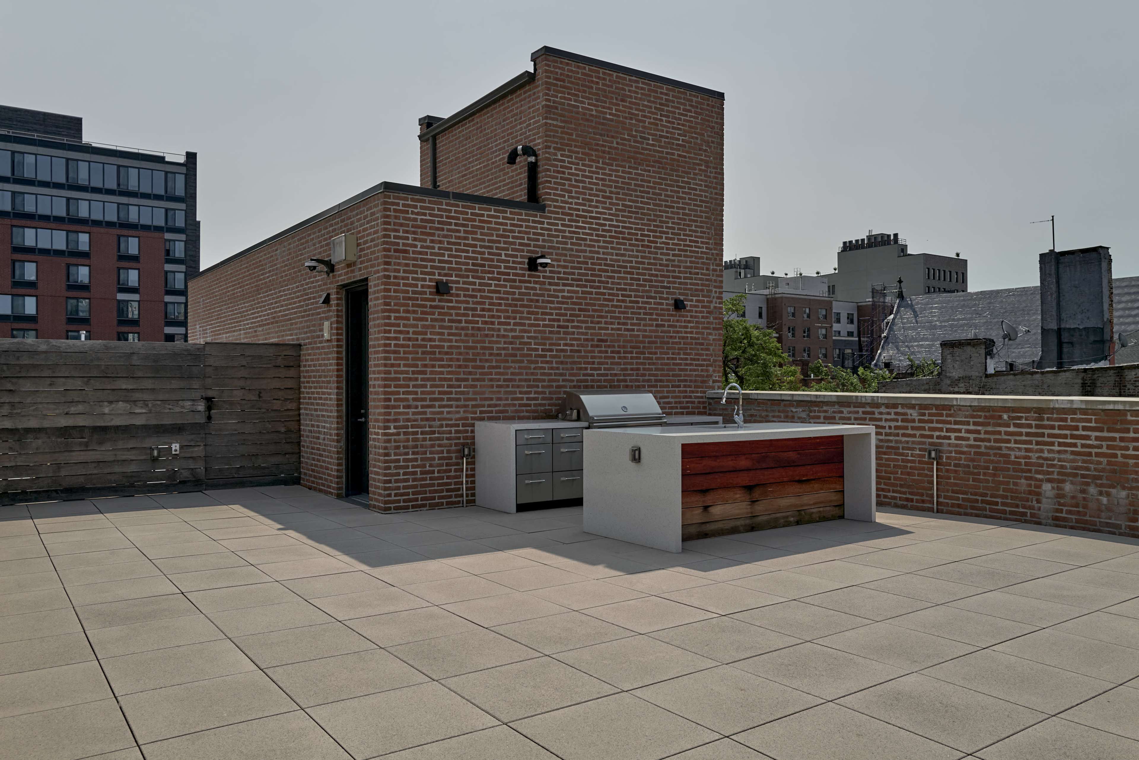 The image shows a rooftop terrace featuring a countertop with a built-in grill and storage, surrounded by brick walls and a clear sky.