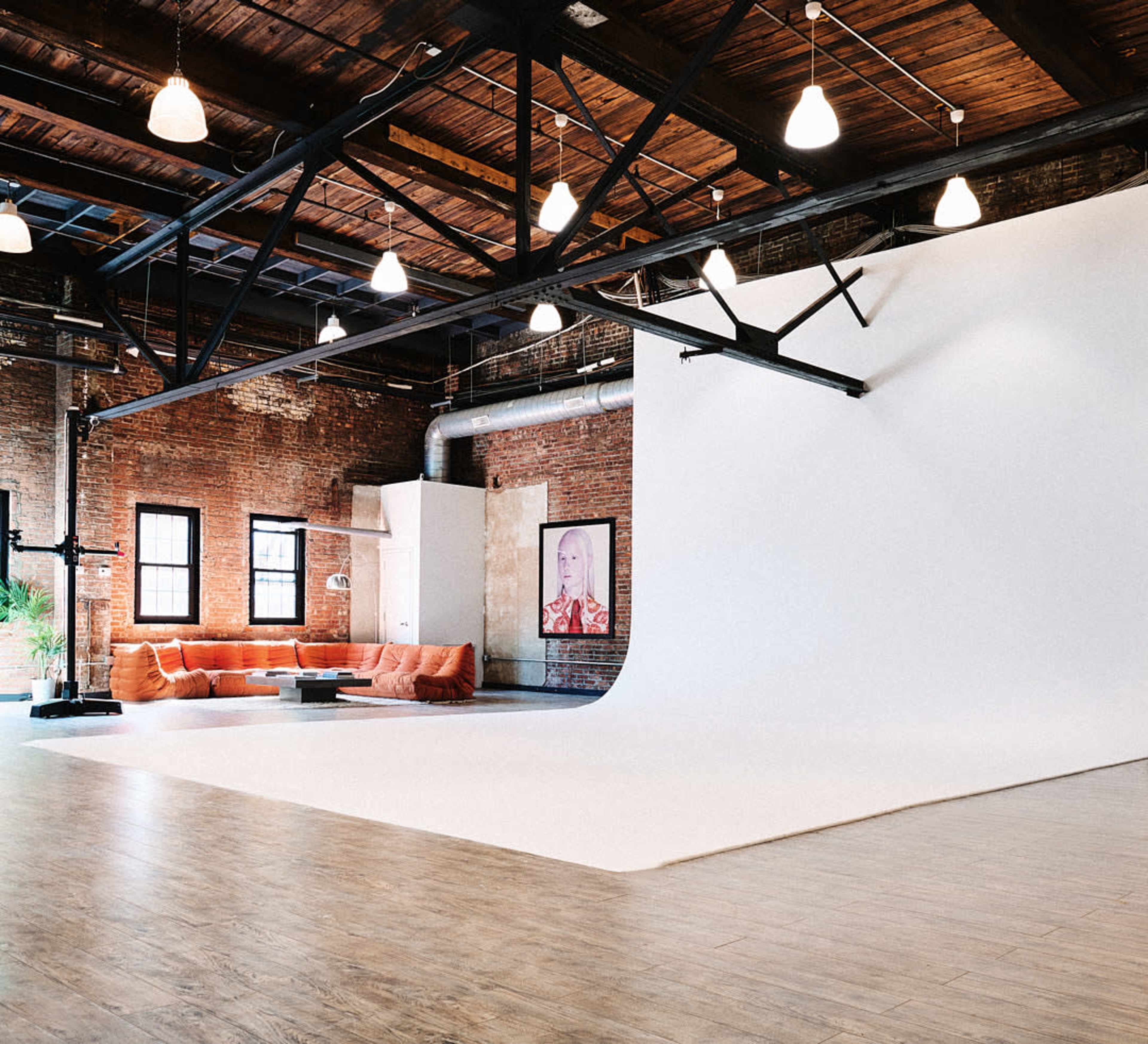 A spacious studio with exposed brick walls, a large white backdrop, and a cozy seating area featuring orange couches.