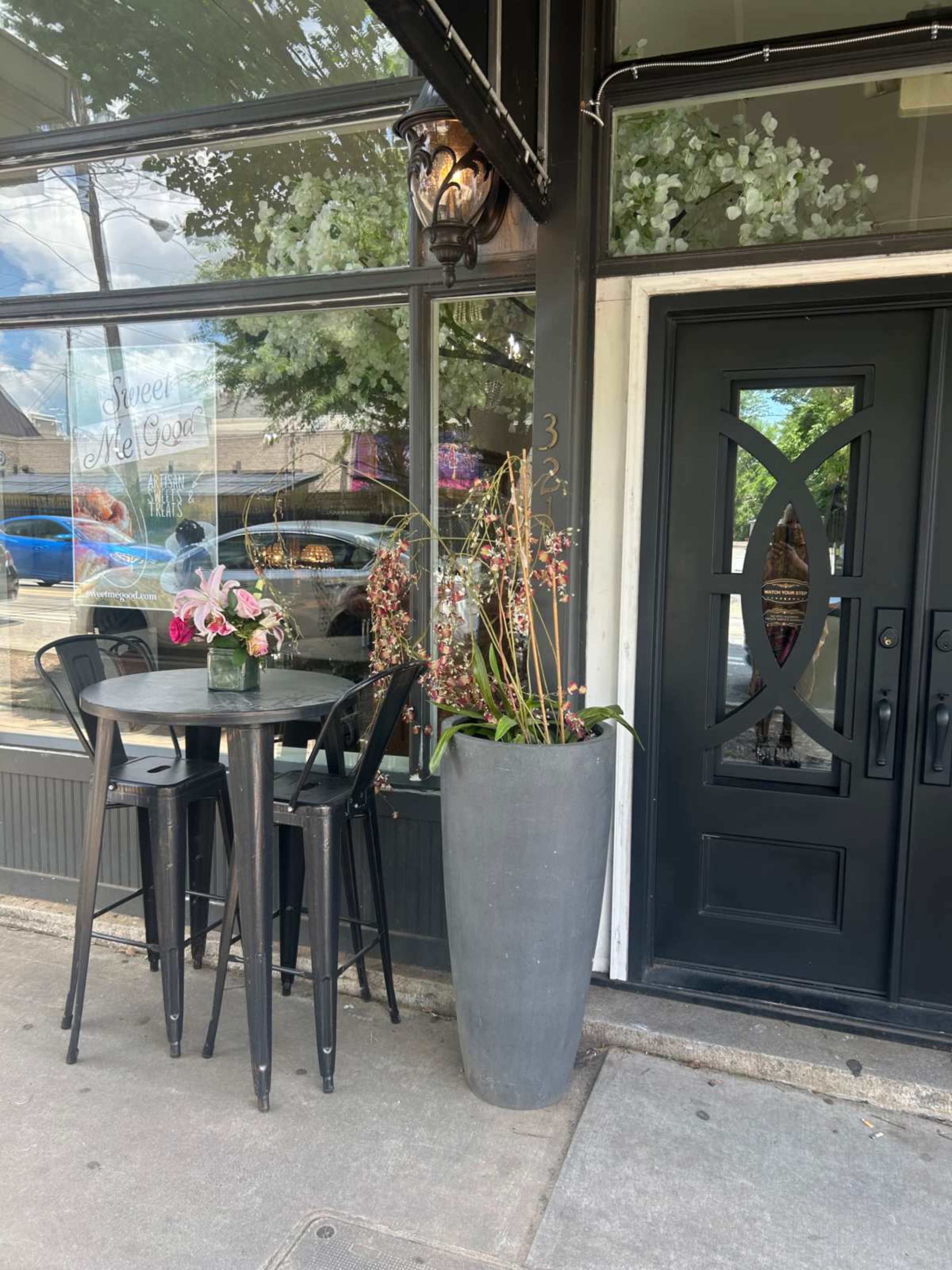 The image shows a small outdoor seating area in front of a restaurant featuring a table with black chairs and a large planter with flowering plants beside an entrance door.