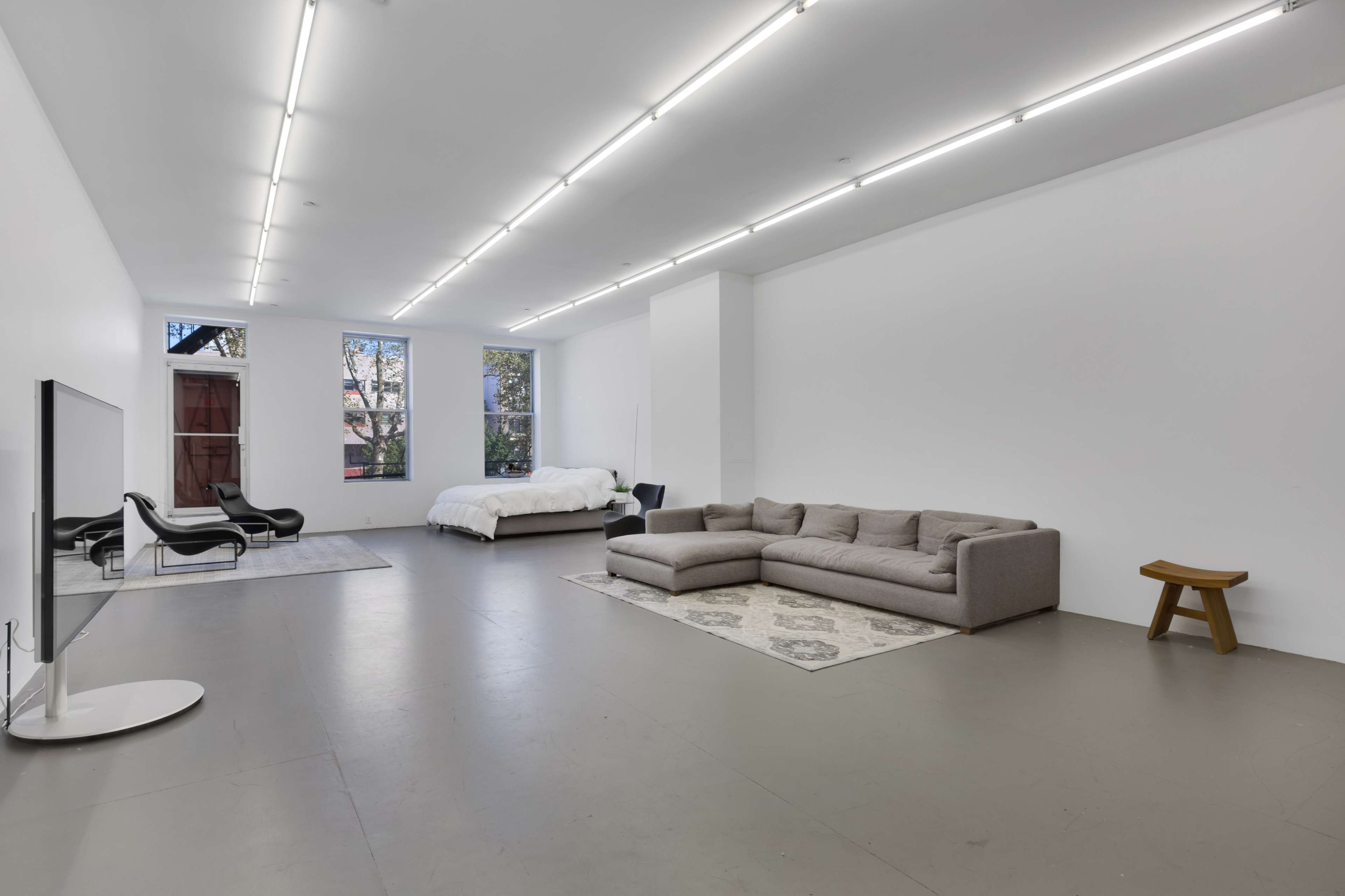 A spacious, modern room featuring a large sectional sofa, a bed, a TV on a stand, and two chairs, all against white walls and a concrete floor.