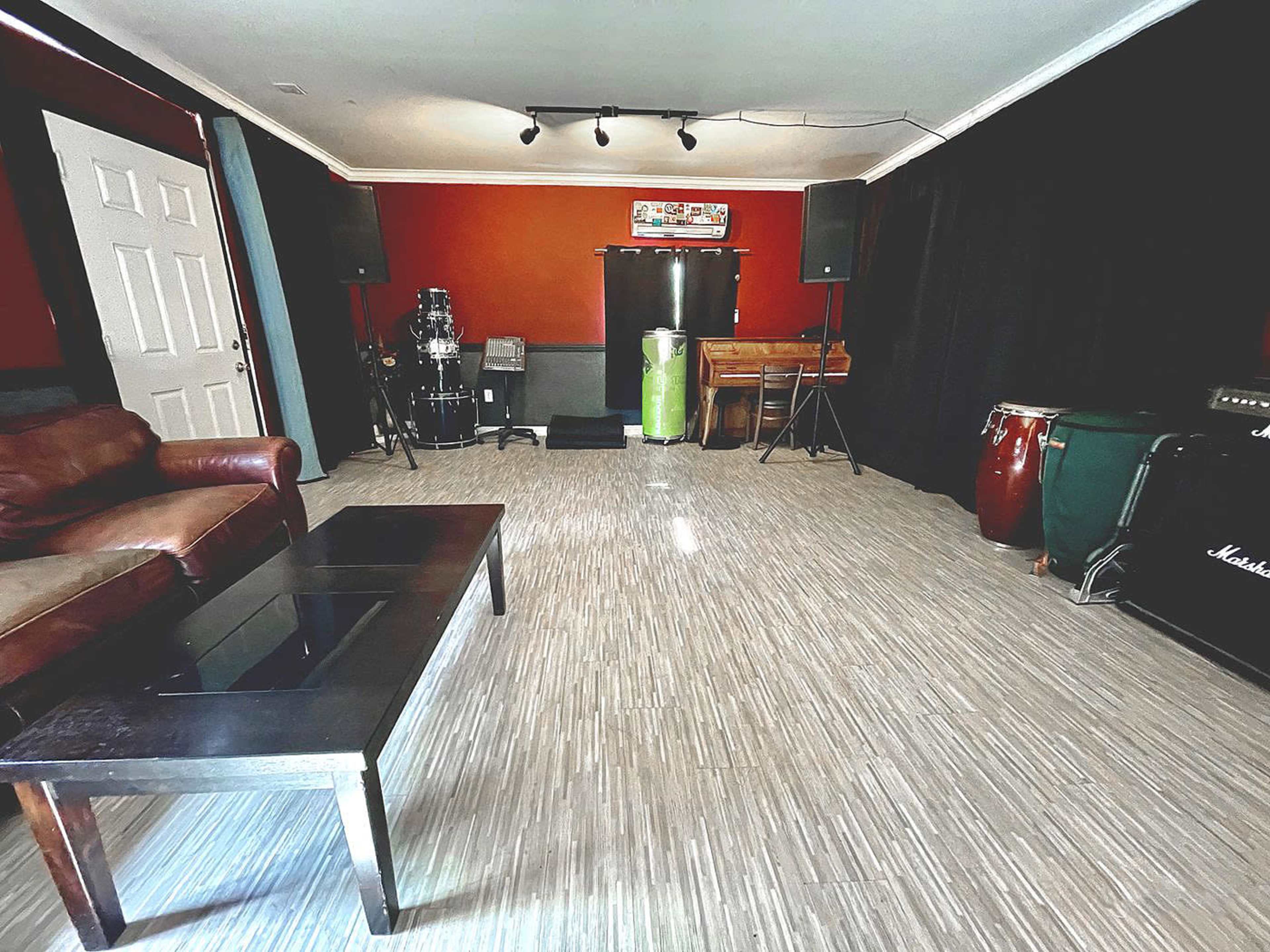 The image shows a music rehearsal space featuring various musical instruments, a leather couch, and a coffee table on a light-colored floor.
