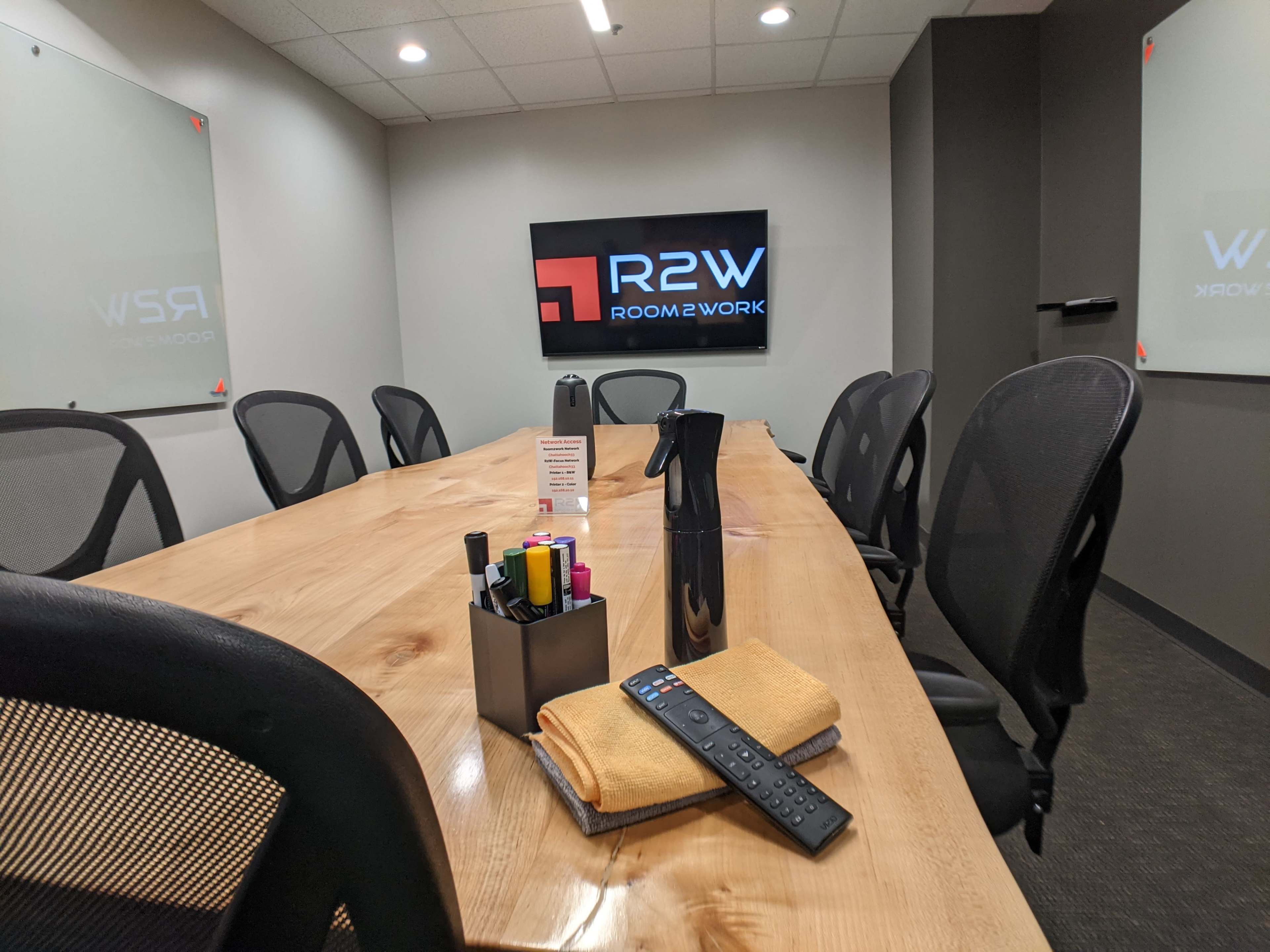 A conference room features a long wooden table with office supplies, a remote control, and a television displaying the logo "R2W Room2Work."