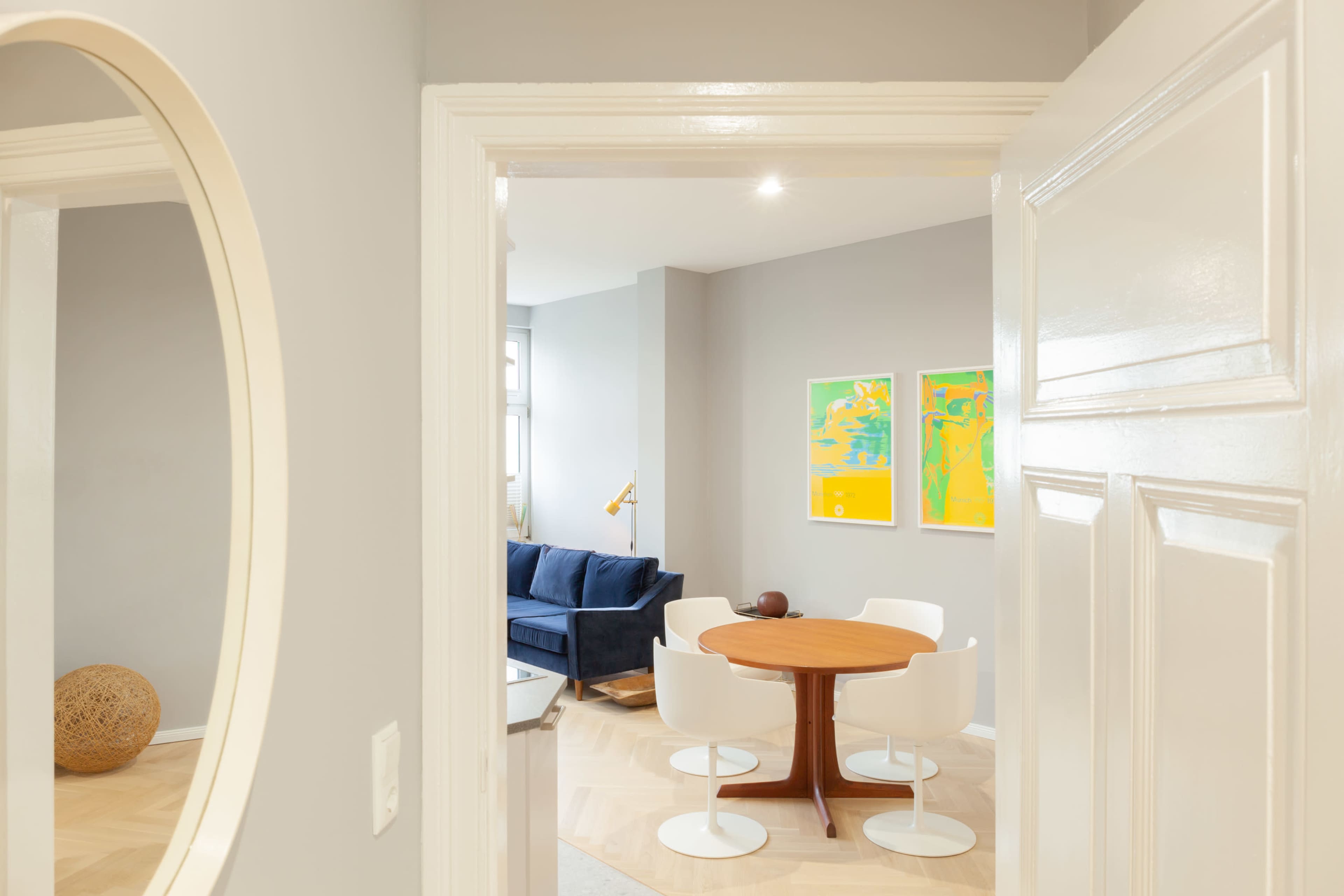 A view through an open door into a room featuring a round wooden dining table with white chairs, a blue sofa, and colorful artwork on the walls.