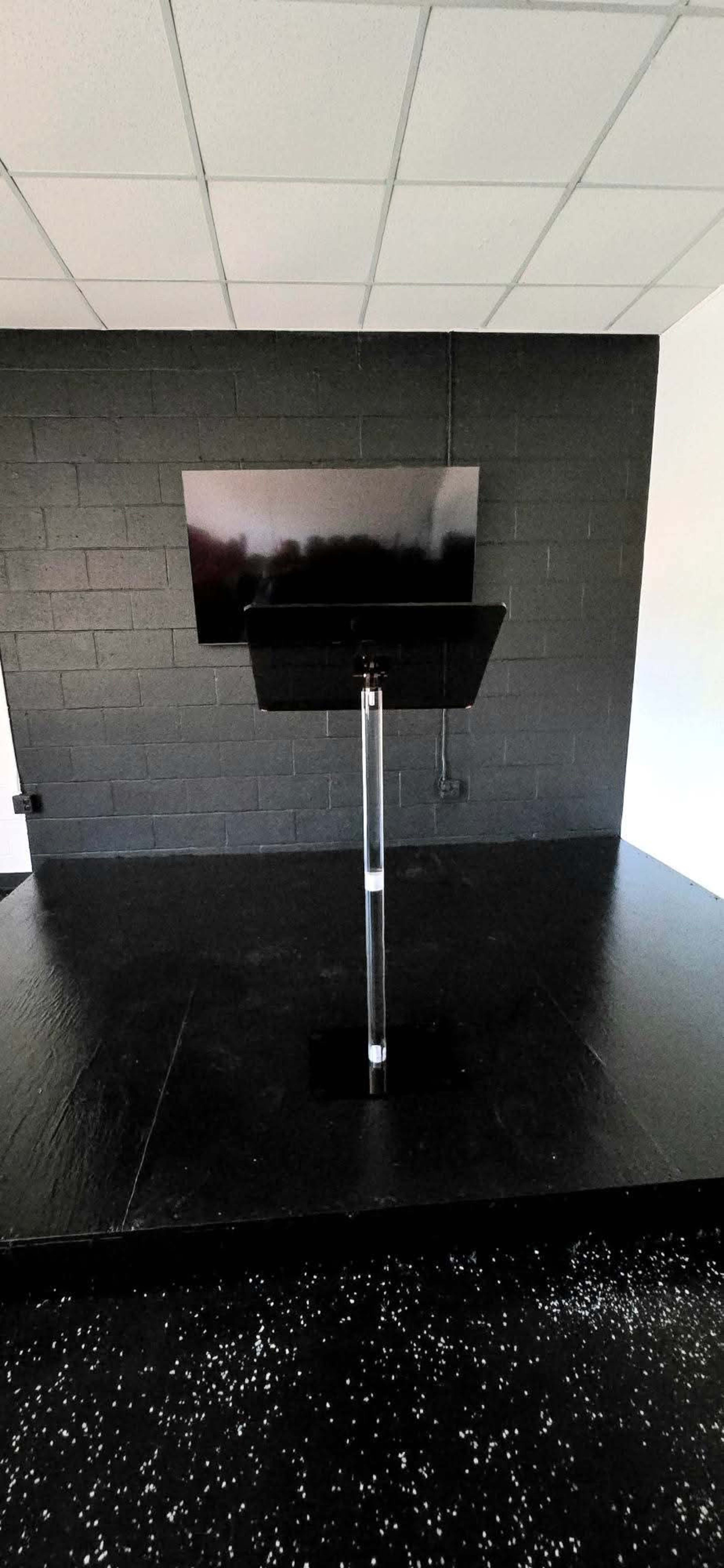 The image shows a clear podium positioned in front of a large black screen against a black wall in a sparsely decorated room.