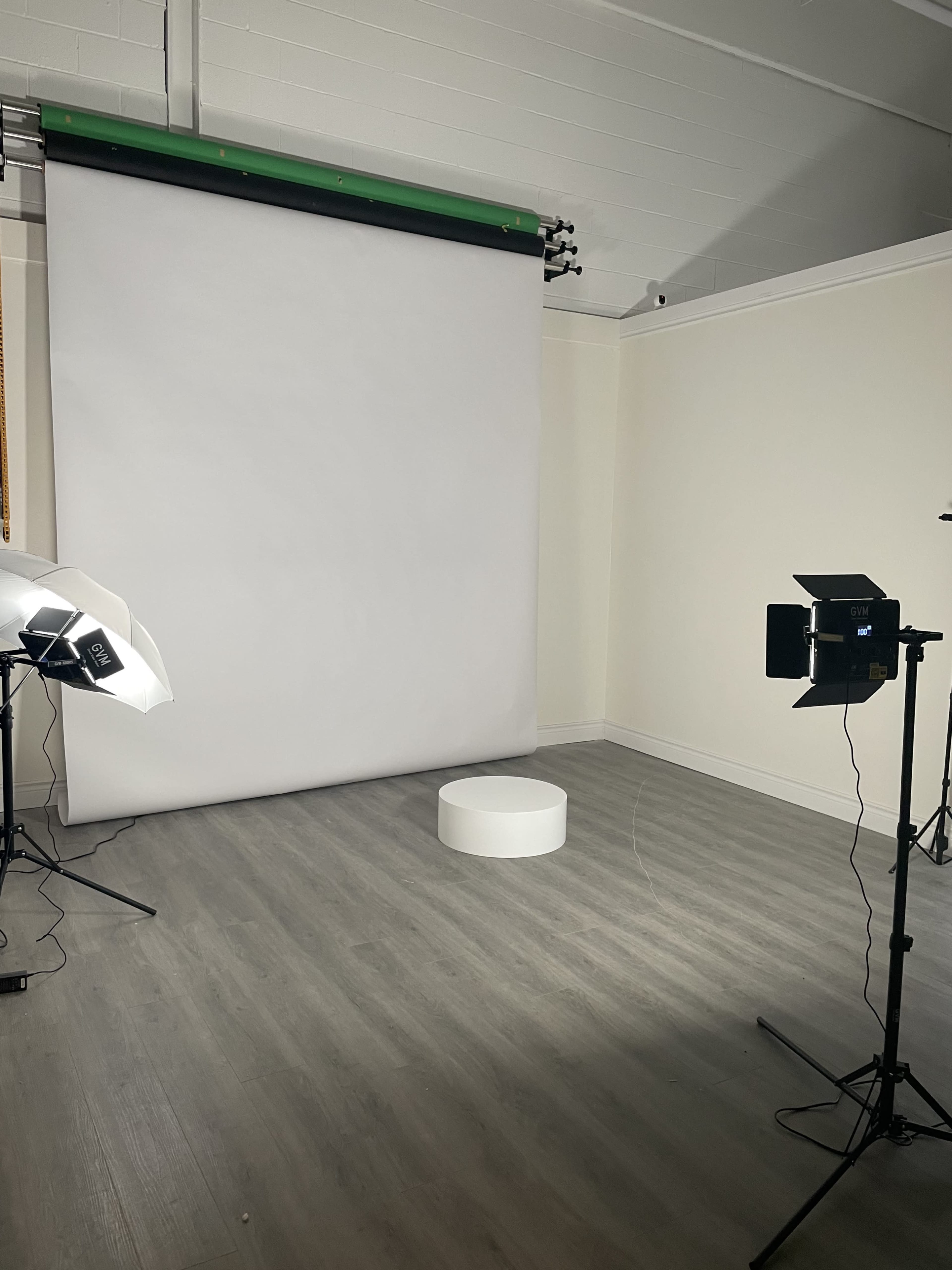 The image shows a photography studio with a white backdrop, a circular platform in the center, and lighting equipment on either side.