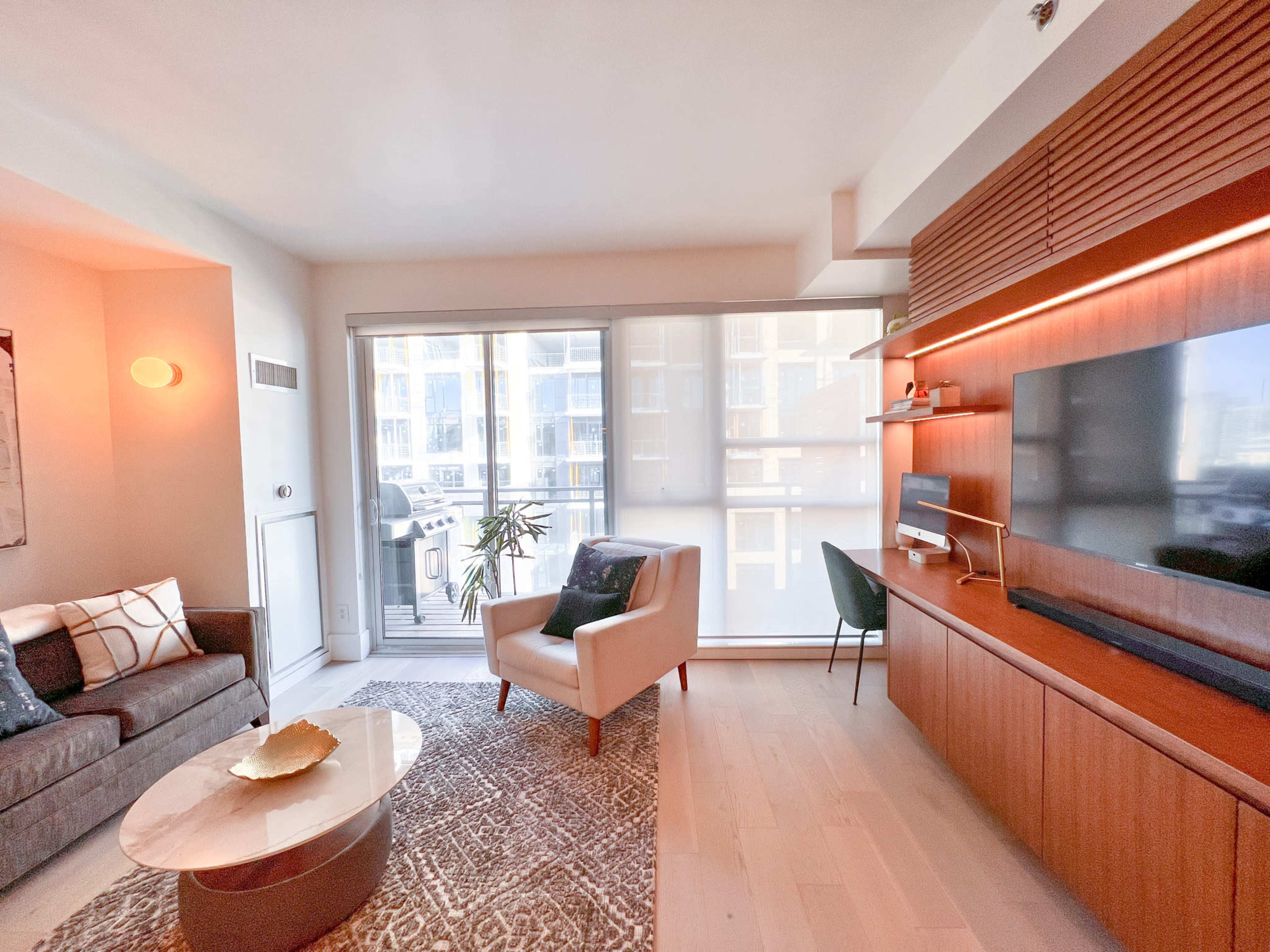 A modern living room featuring a gray sofa, a light-colored armchair, a round coffee table, and a wall-mounted television with wooden cabinetry, all enhanced by large windows letting in natural light.