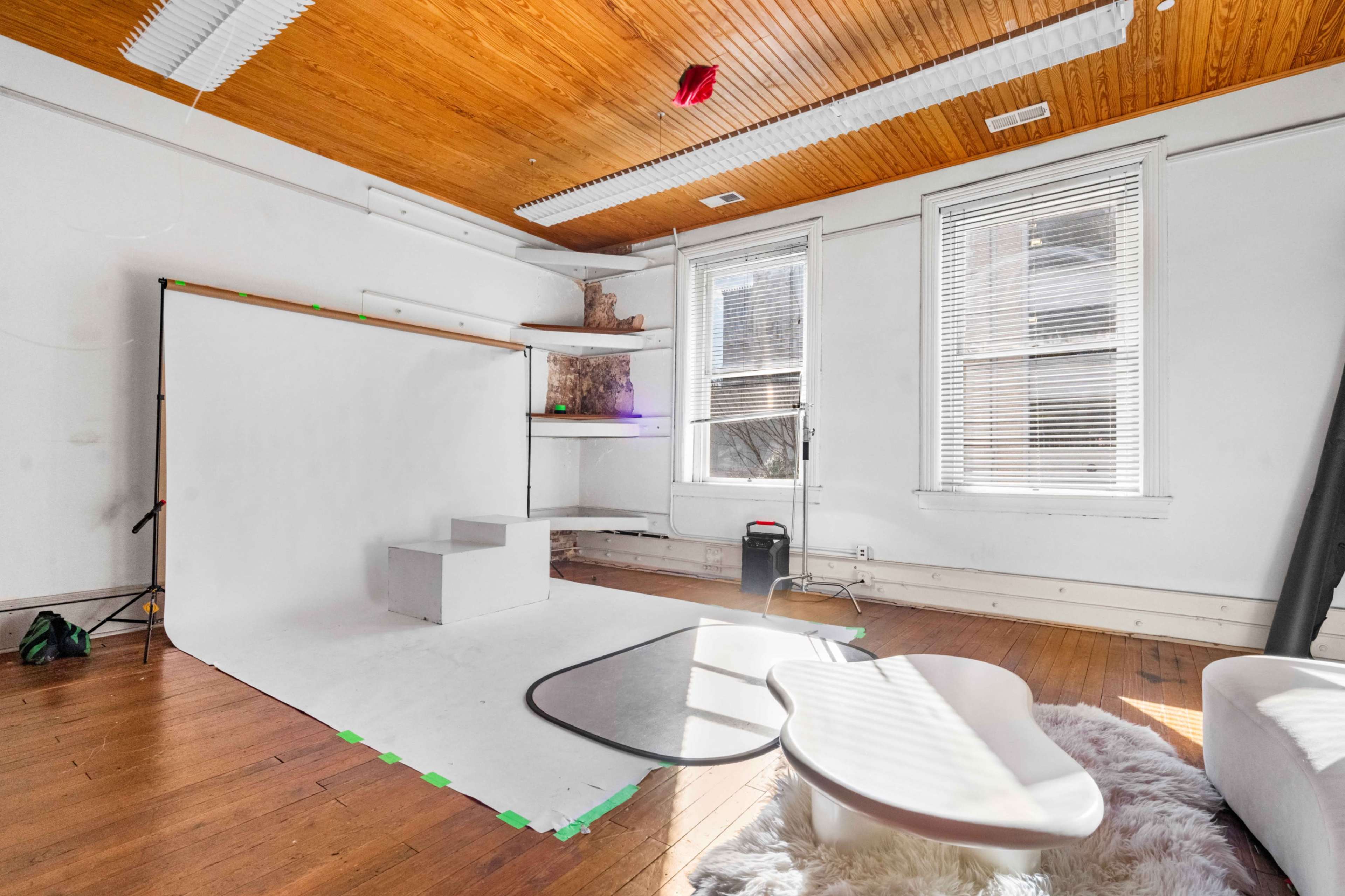 The image shows a bright, empty studio space with a white backdrop, a small raised platform, and natural light coming through large windows.