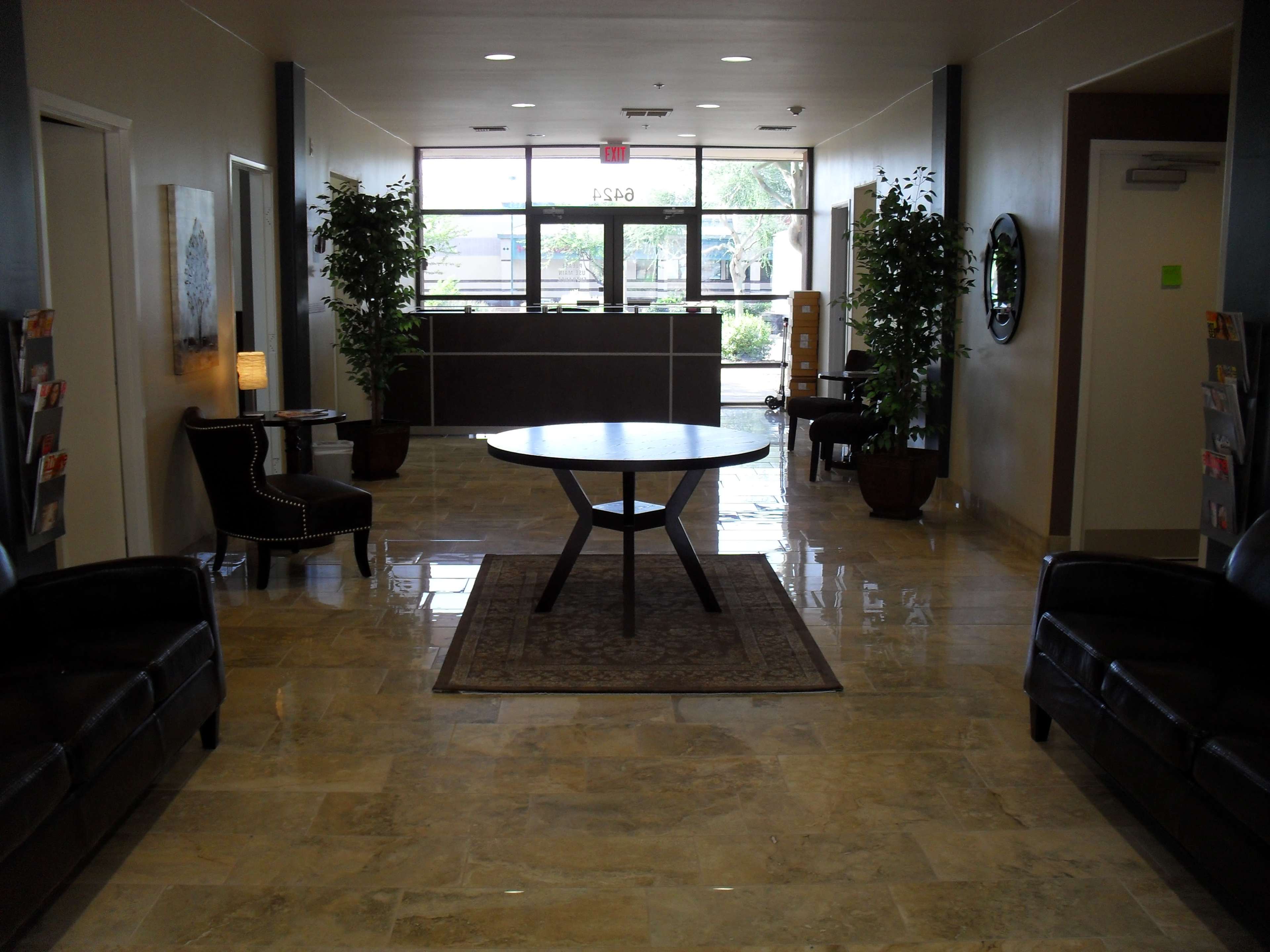 A modern lobby with a circular table, two leather chairs, and plants lining the sides, leading to a reception area with large windows.