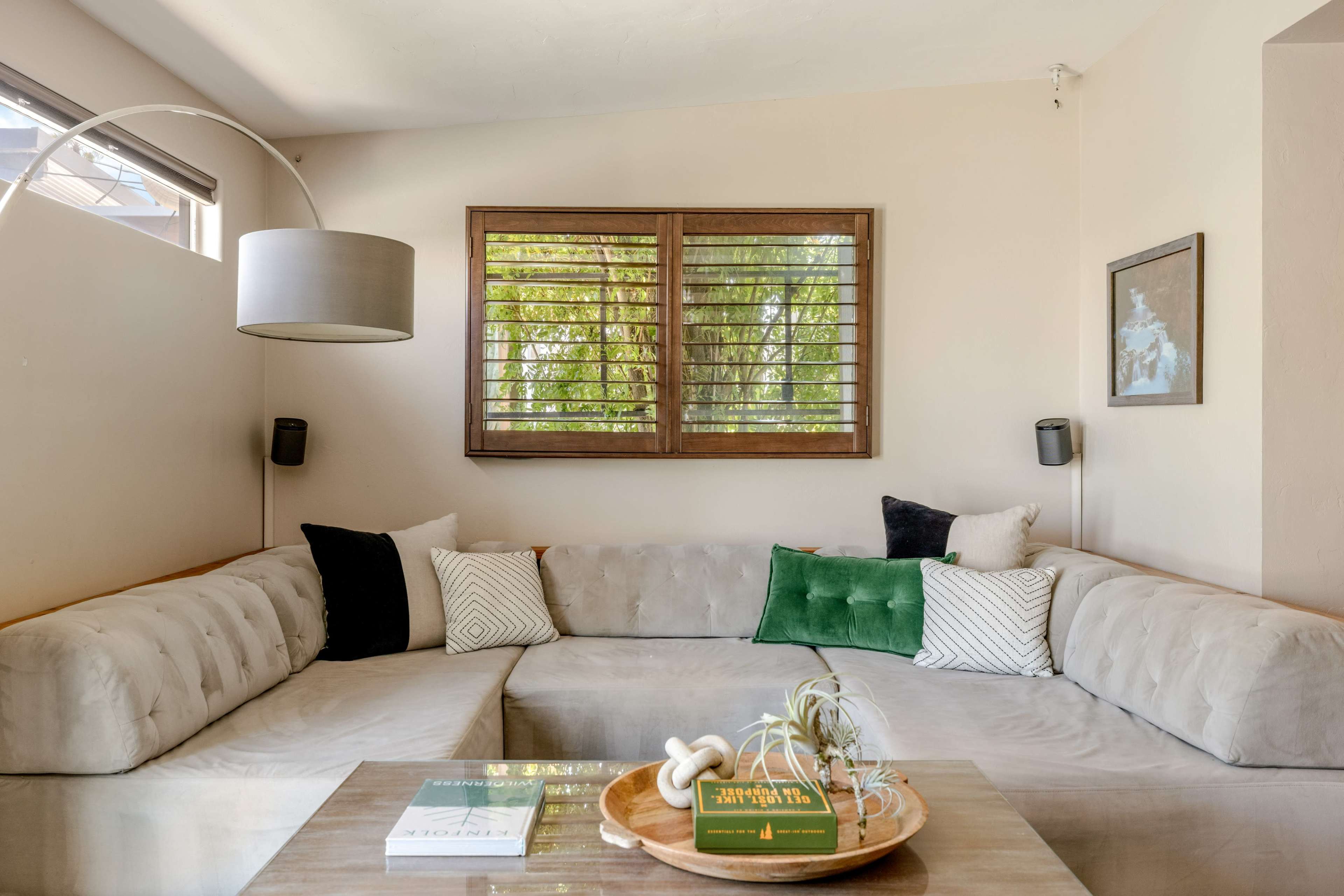 The image shows a modern living room with a light-colored, U-shaped sofa, wooden window shutters, and a coffee table featuring decorative items and plants.