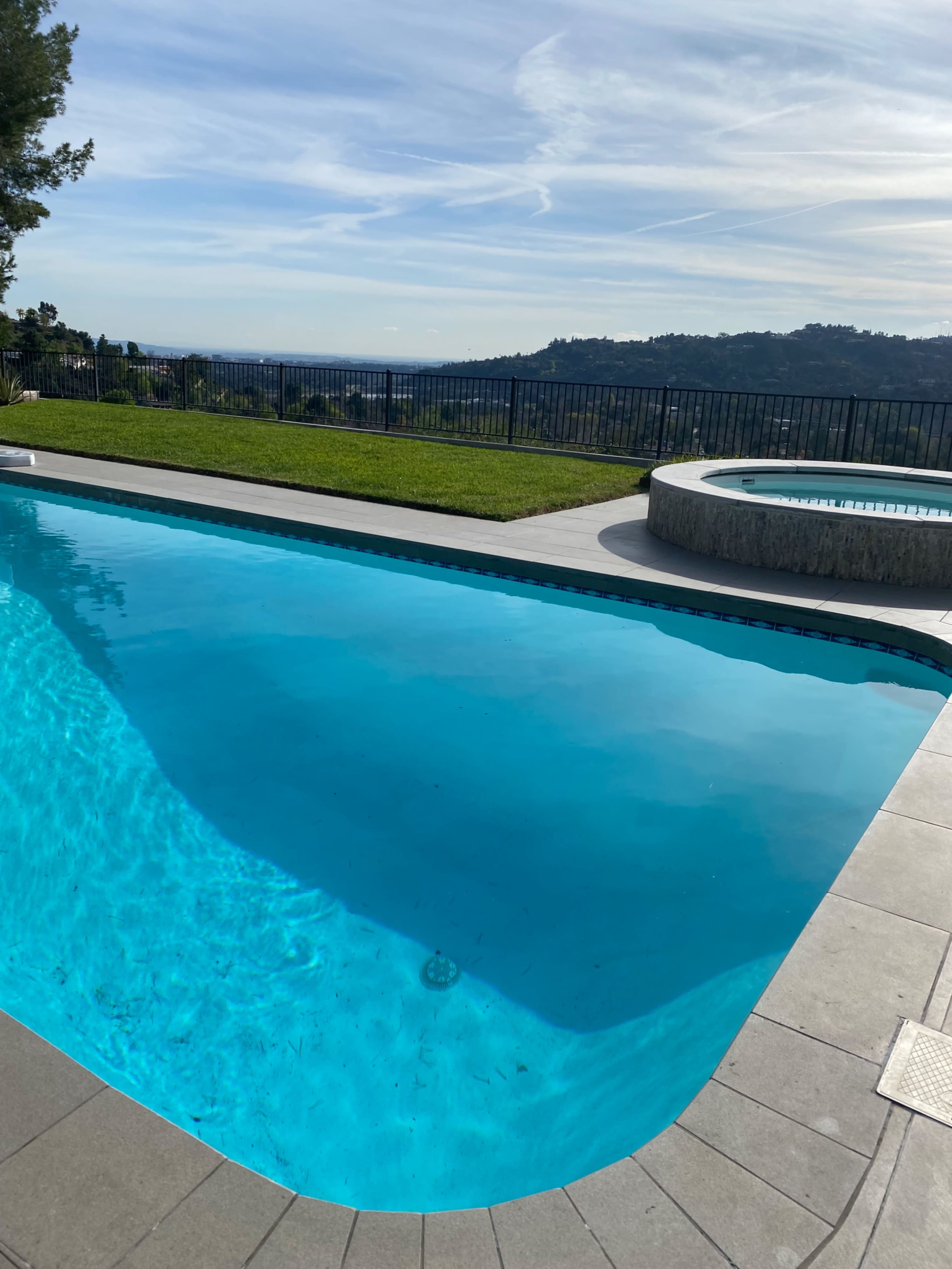 A clear swimming pool with a circular hot tub is set against a scenic hillside view.