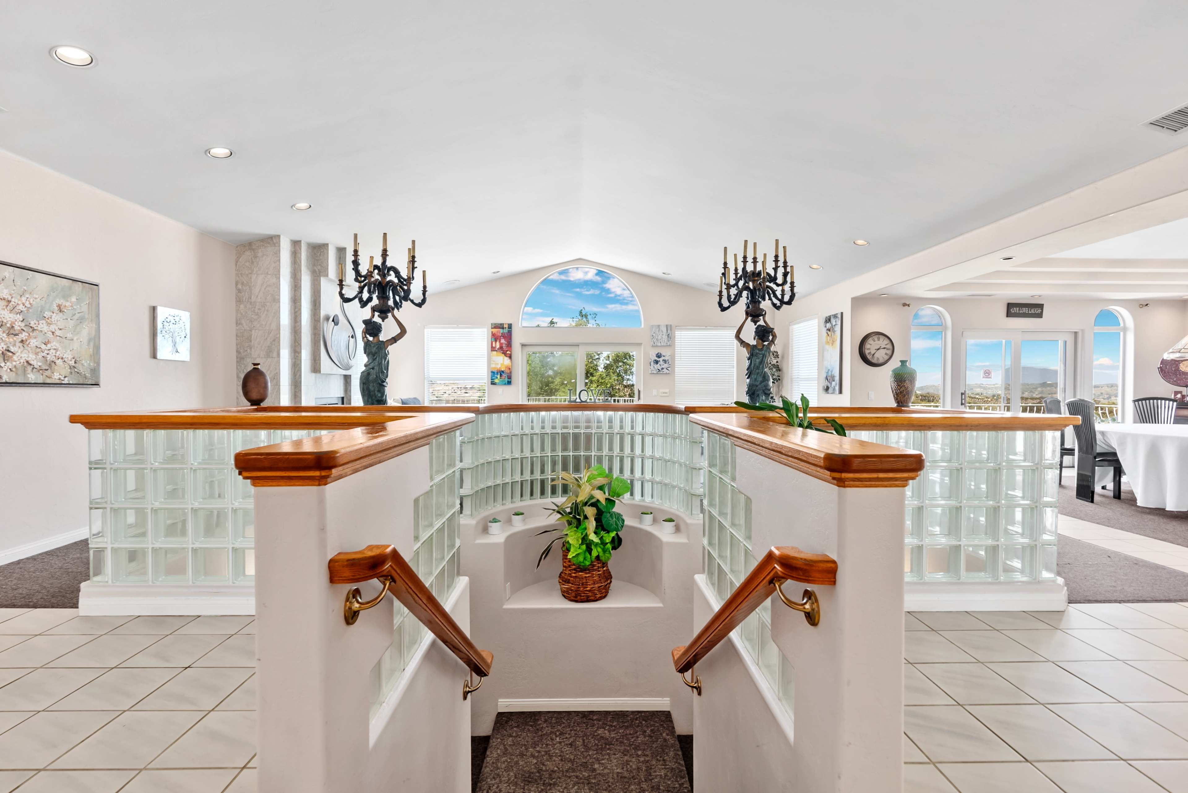 A spacious interior featuring a curved staircase with glass block railings, potted plants, and large windows providing a view of the sky.