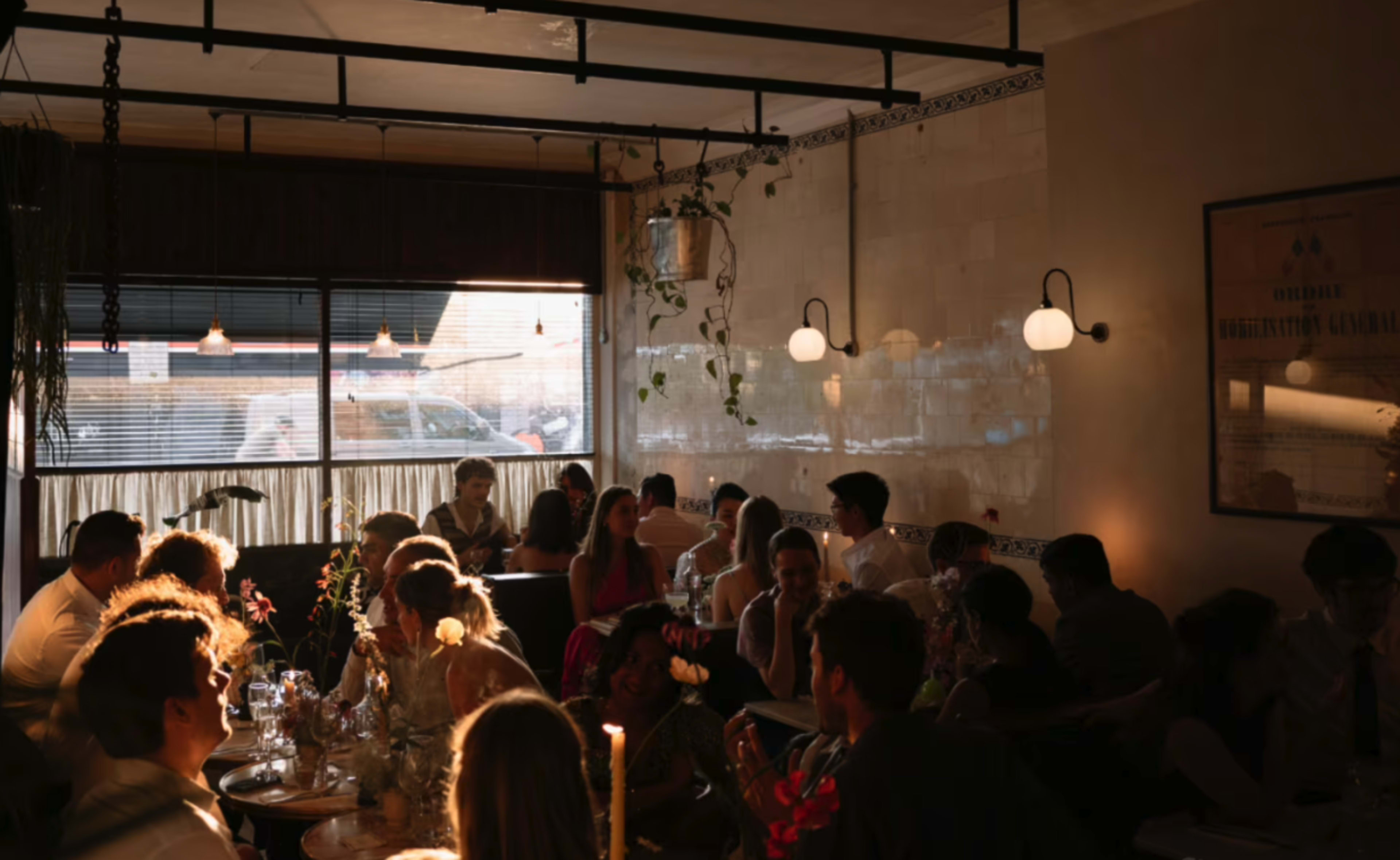 A lively restaurant scene at dusk features patrons gathered around tables, with warm lighting and plants adding to the ambiance.
