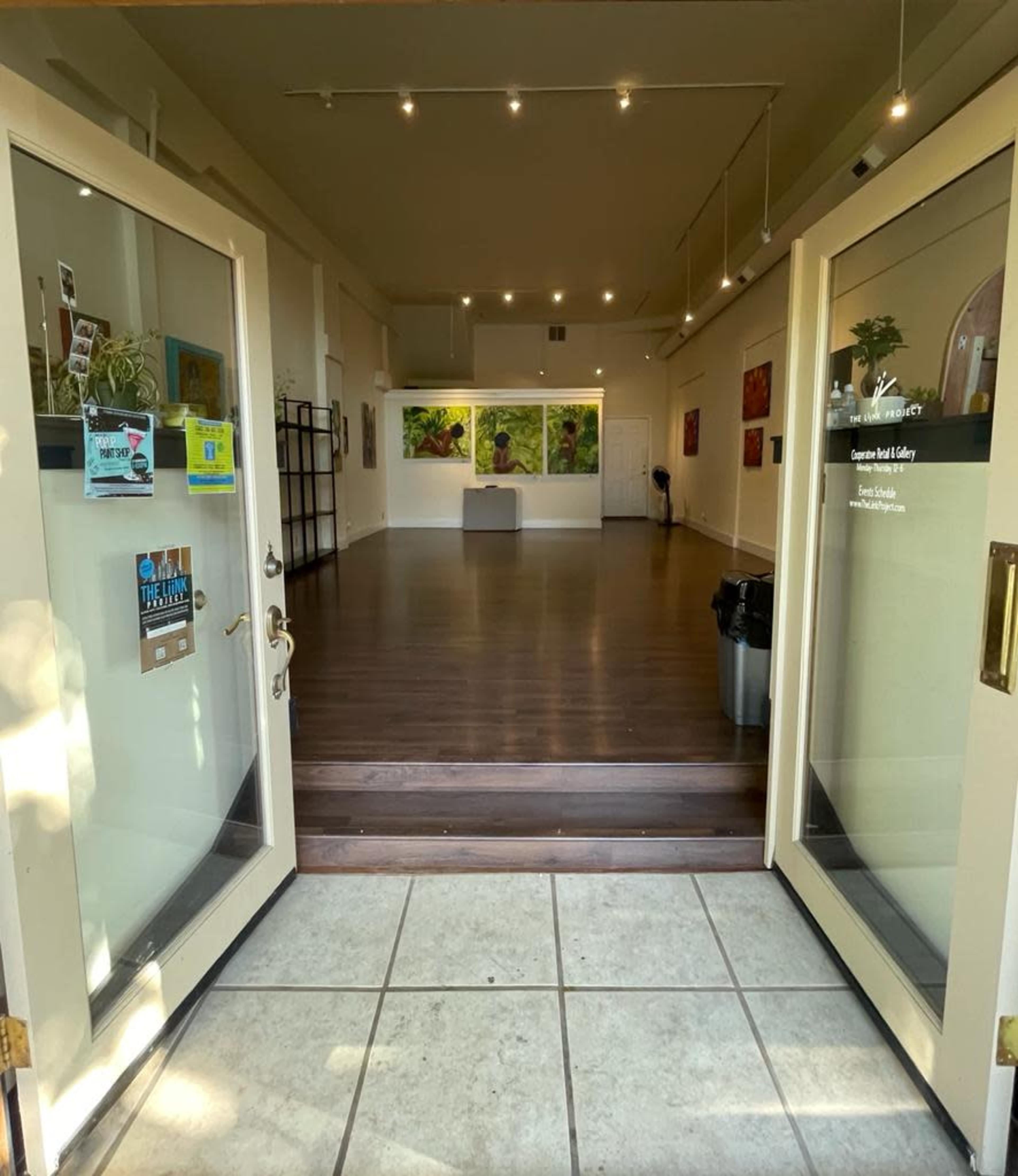 A spacious gallery with open double doors leading into a room featuring wooden flooring and artwork on the walls.