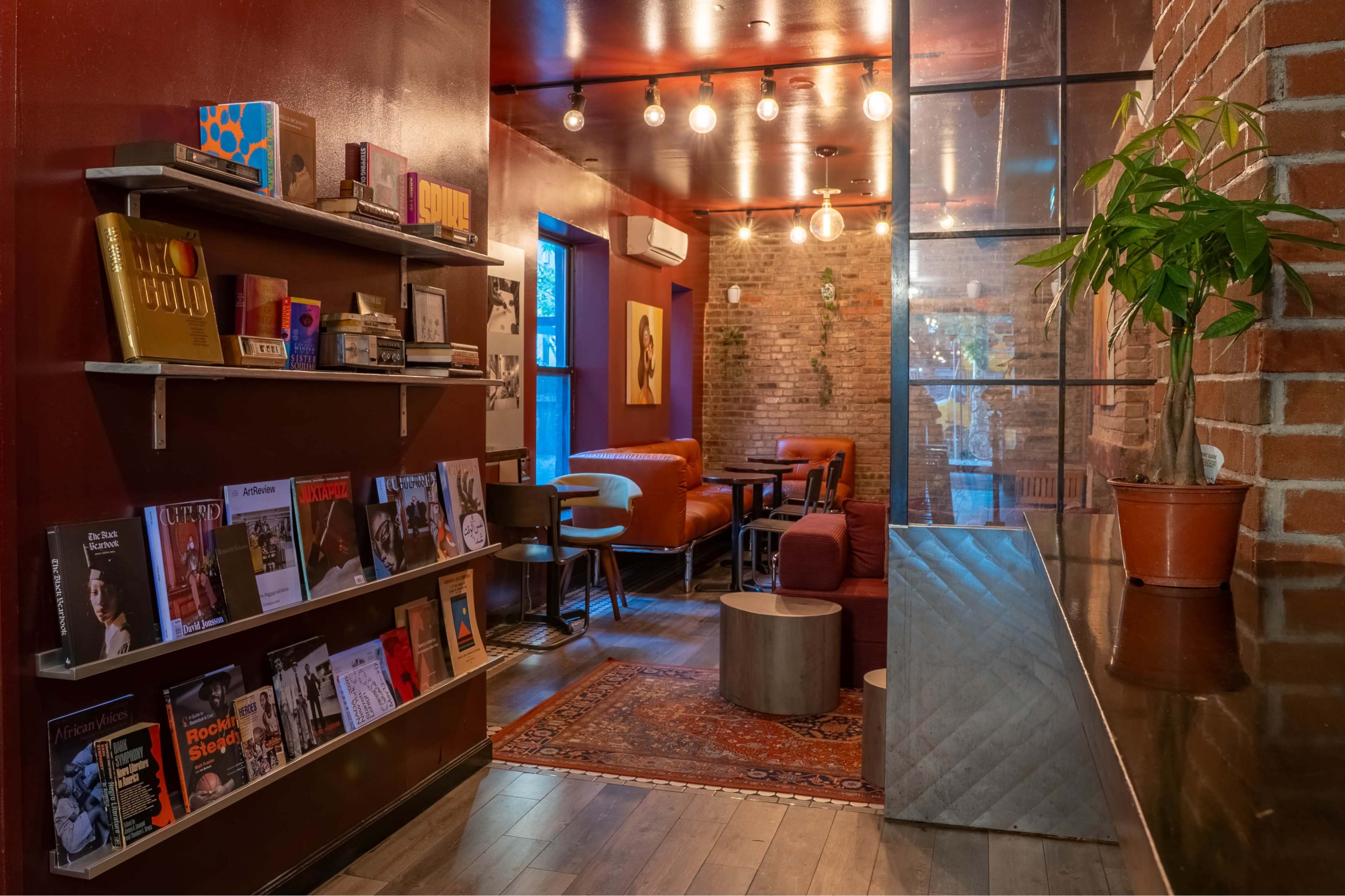 The image shows a cozy interior of a café featuring exposed brick walls, shelves filled with books and magazines, and warm lighting from hanging bulbs.