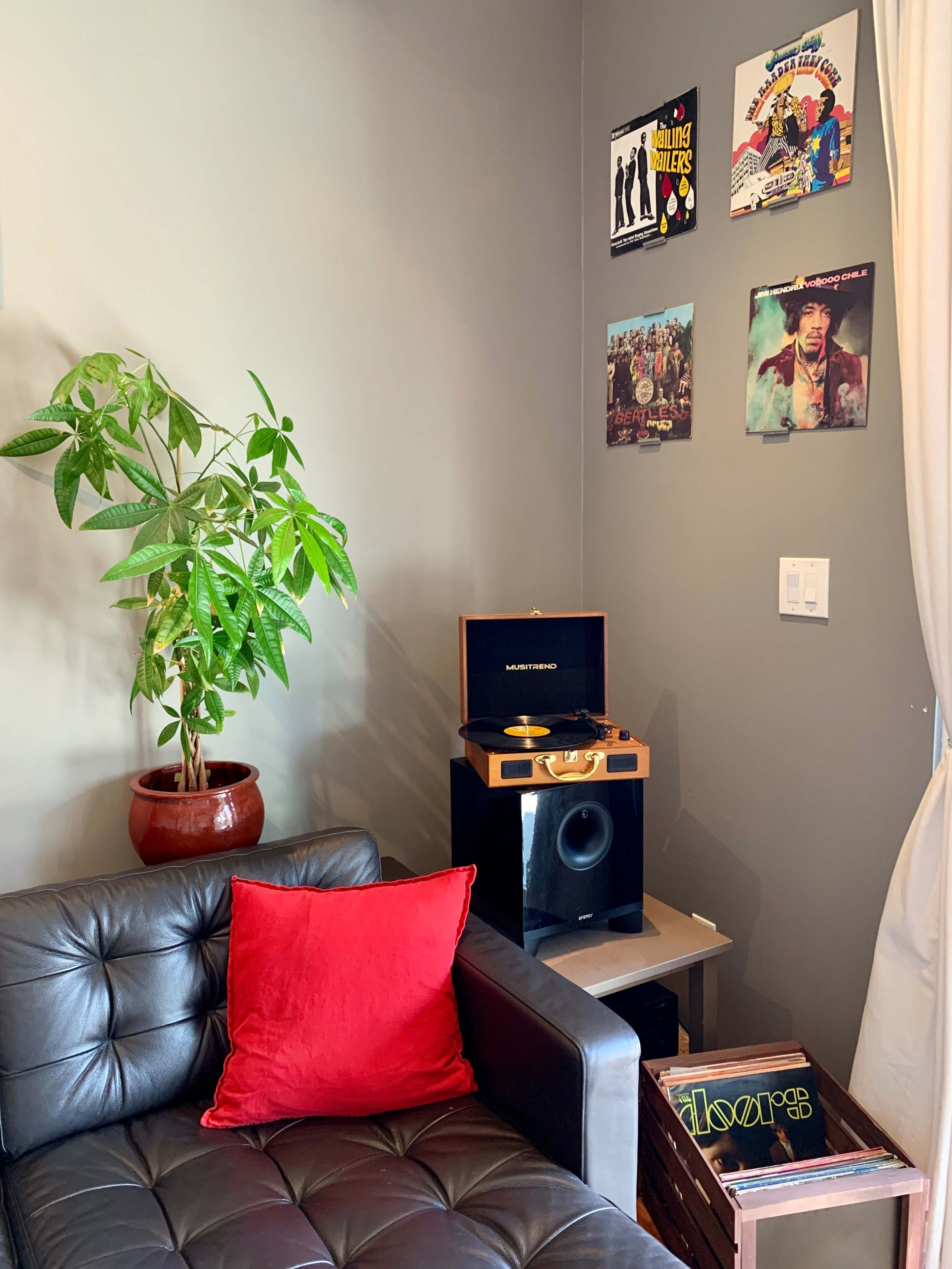 A black leather couch with a red pillow sits beside a record player, potted plant, and framed vinyl records on the wall.