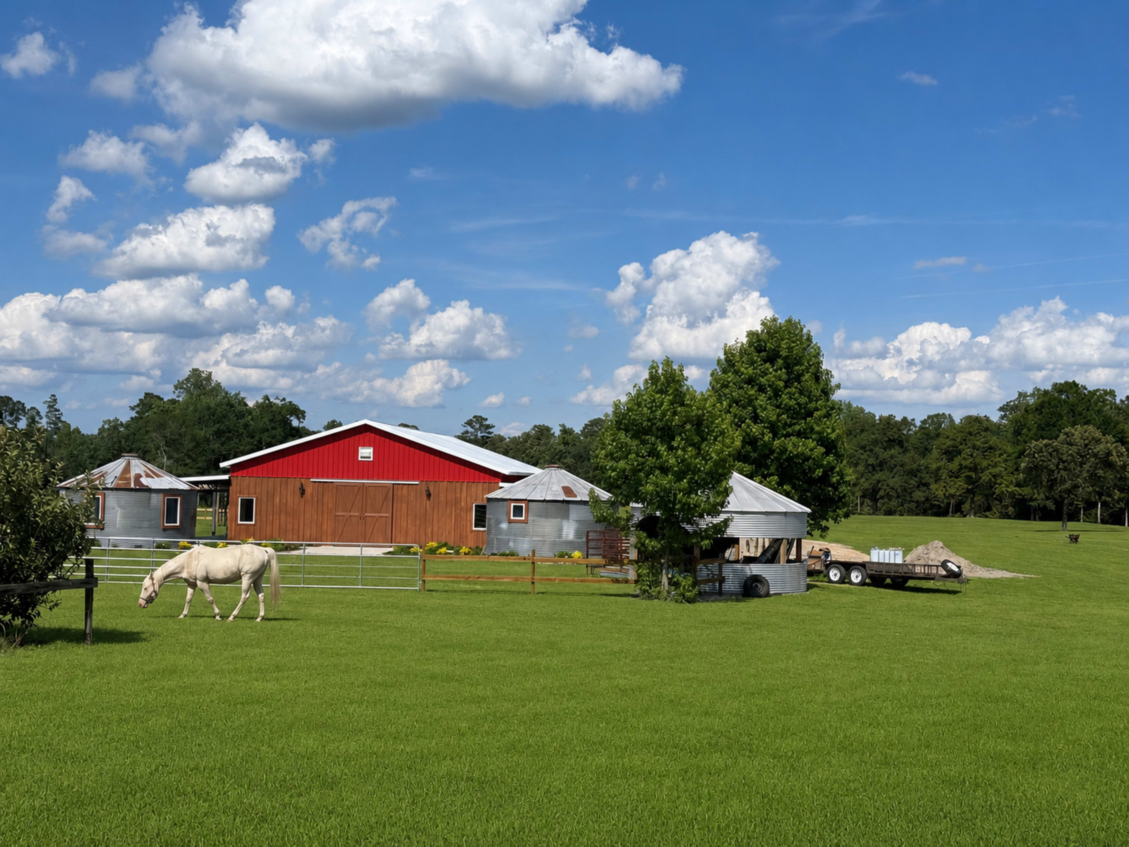A white horse grazes in a green pasture next to a red barn with silver outbuildings and a trailer parked nearby.