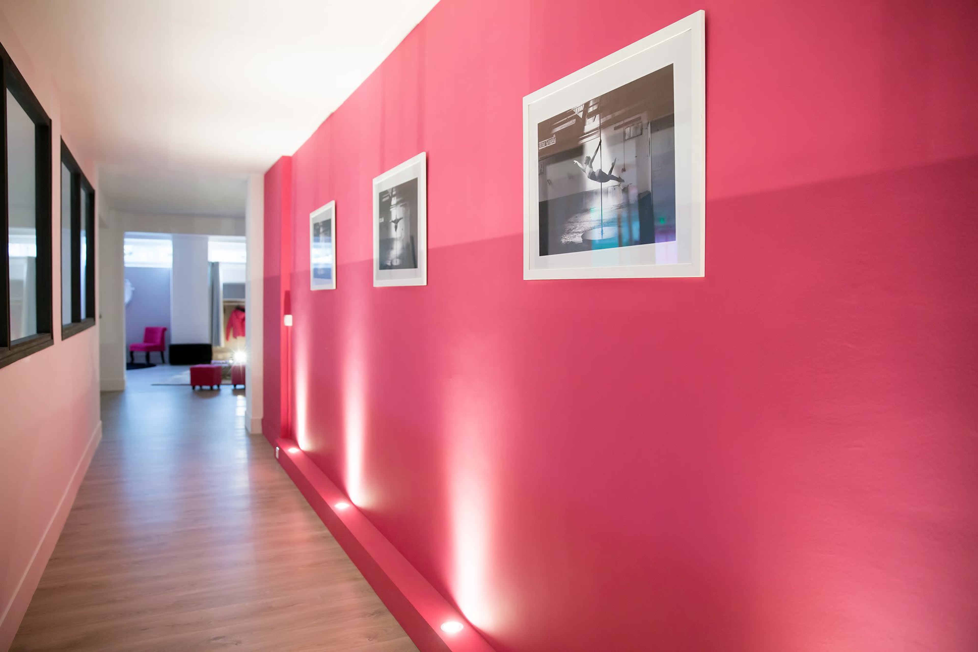 A corridor with pink walls features framed photographs and subtle lighting along the floor.