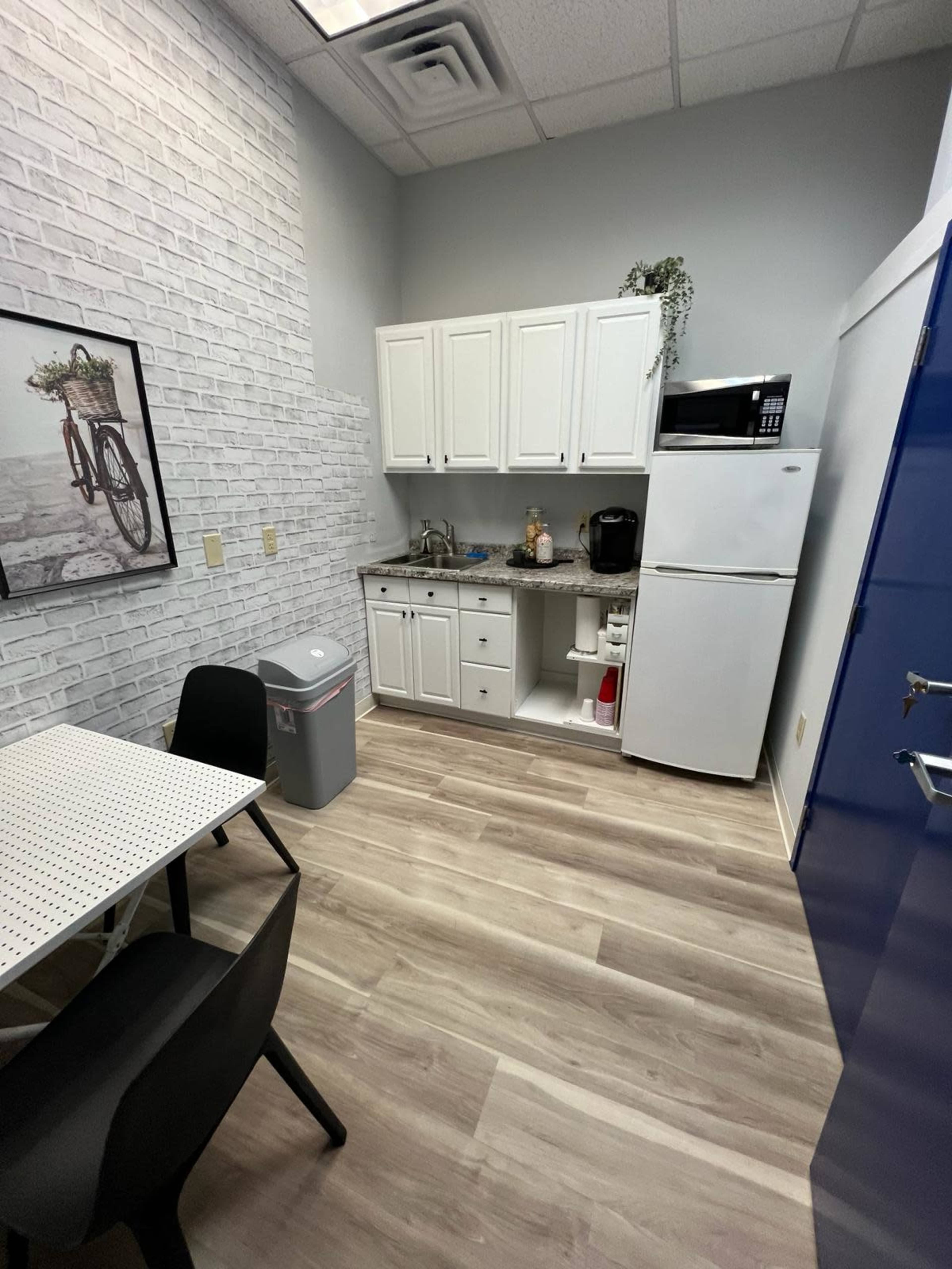 A small kitchen area with a white brick wall, a table with black chairs, and a kitchenette featuring a sink, microwave, and refrigerator.