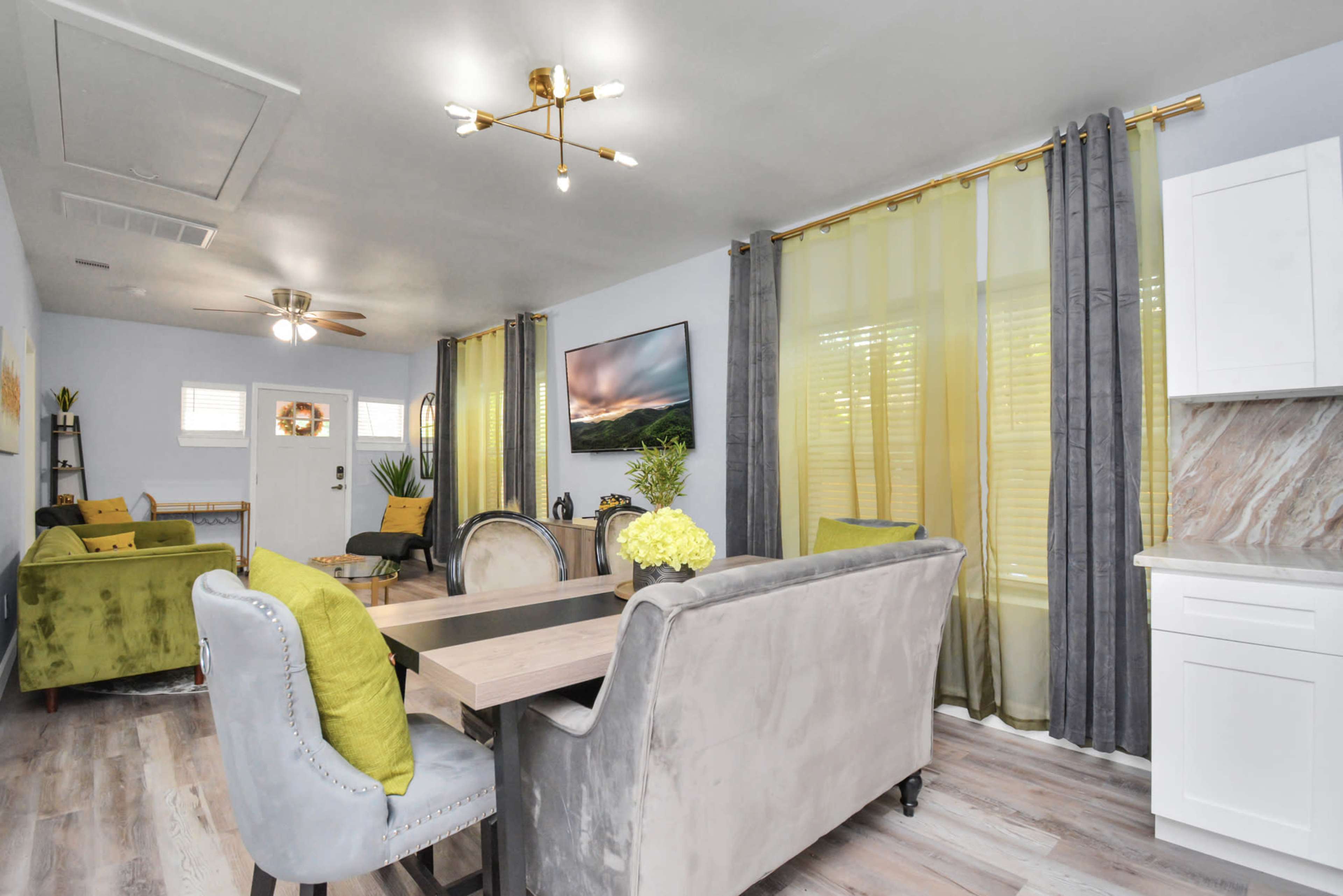 A modern living space features a dining area with a large table, surrounded by upholstered chairs, and a cozy seating area set against large windows dressed in sheer yellow and gray curtains.