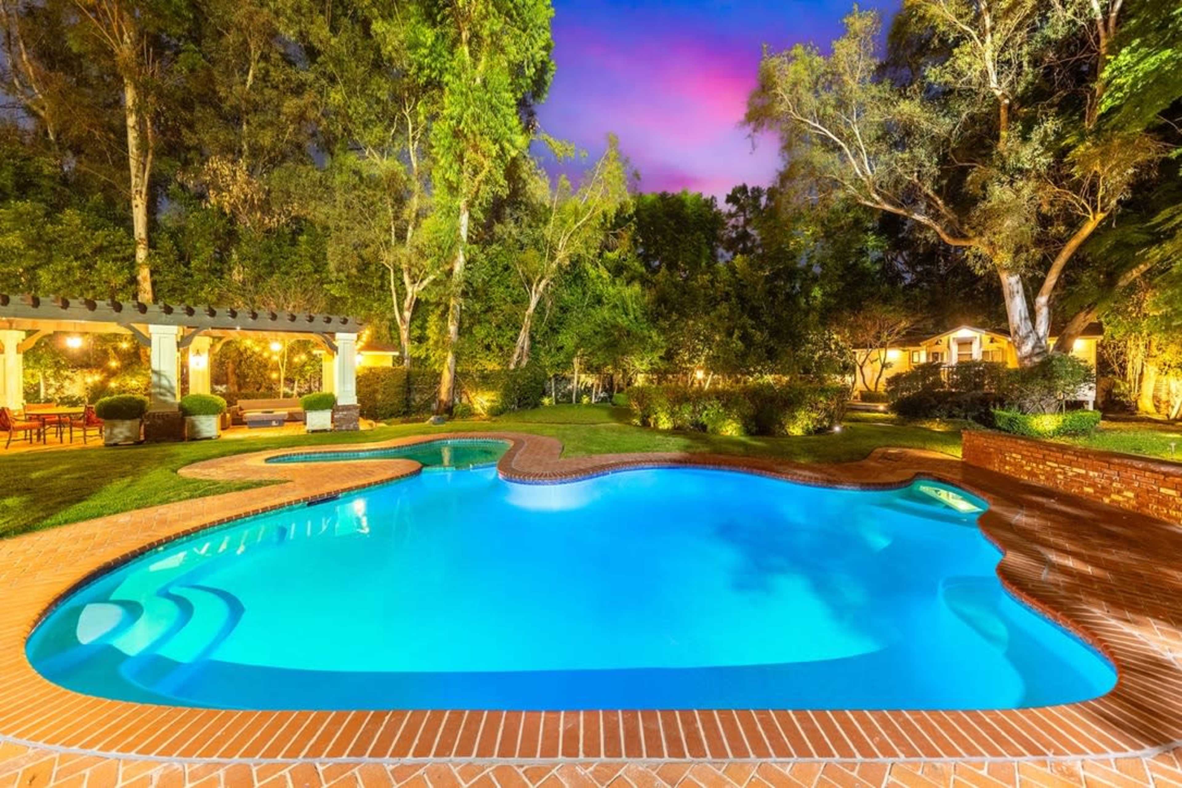 The image shows a well-lit swimming pool surrounded by lush greenery and a patio area at dusk.