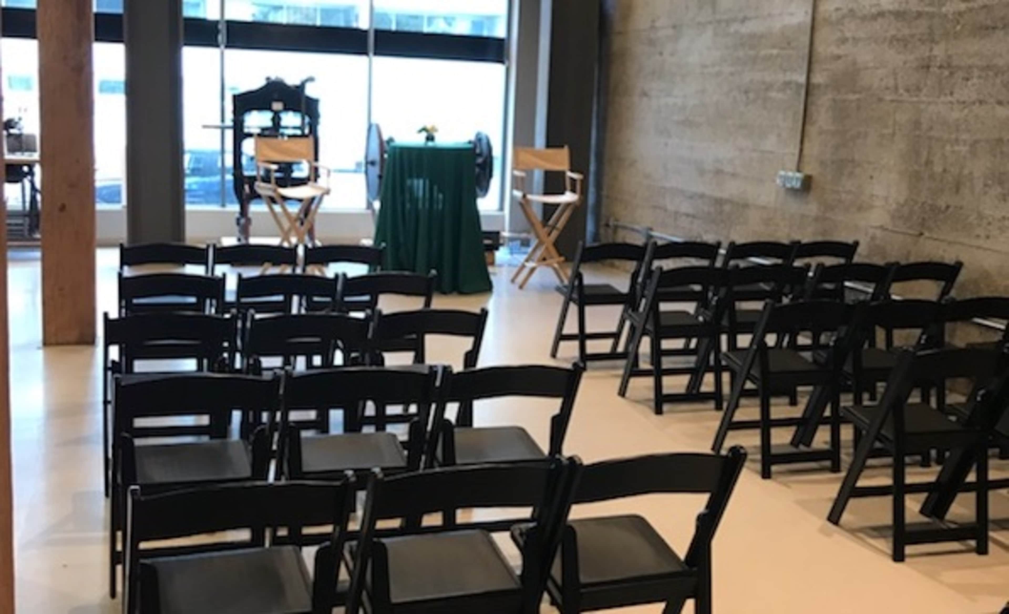 A row of black folding chairs is arranged in front of a green table and two director's chairs in a minimalist room with a concrete wall.