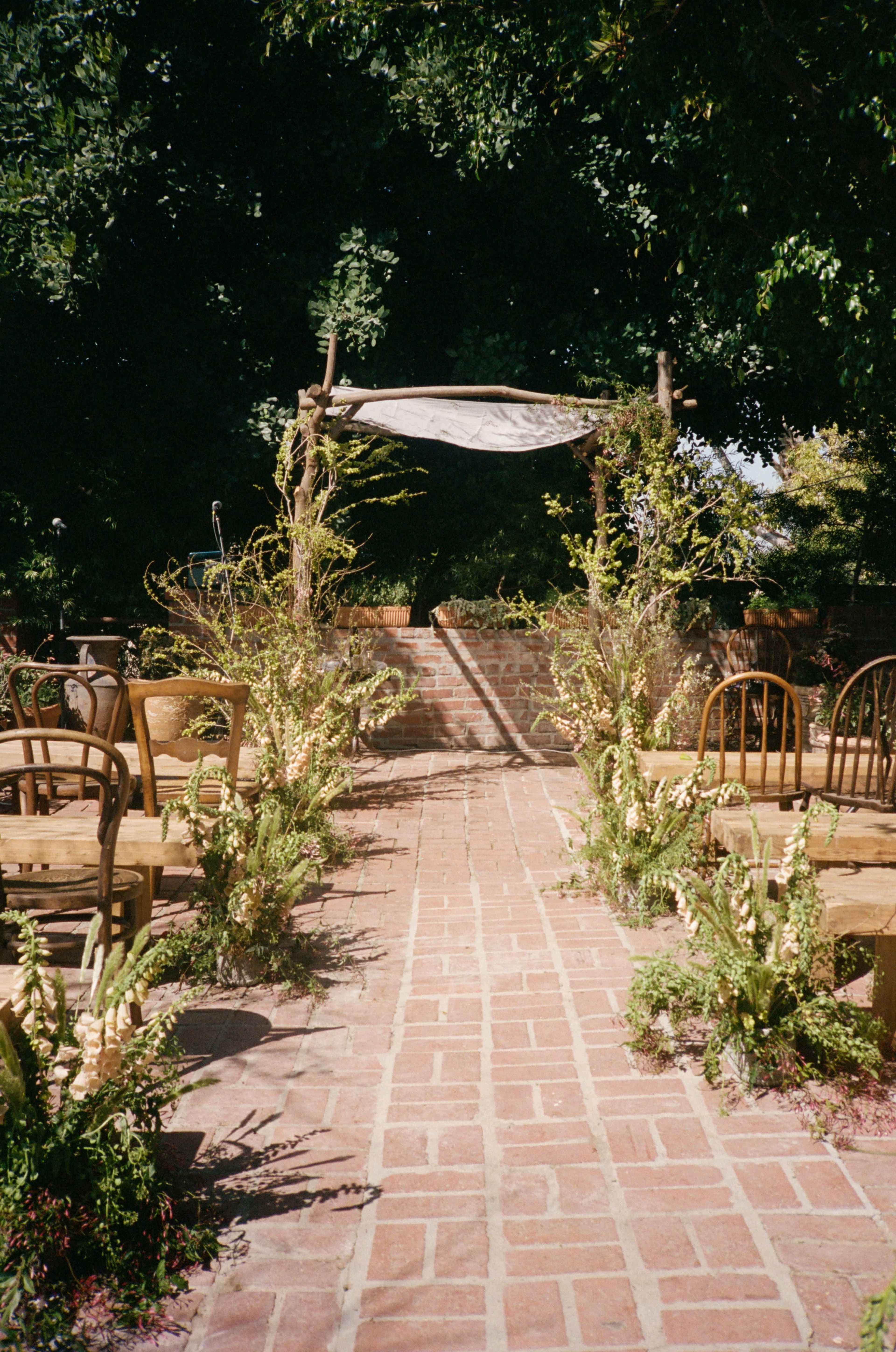 The image shows a brick pathway leading to an open-air ceremony space adorned with wooden chairs and floral arrangements on either side.