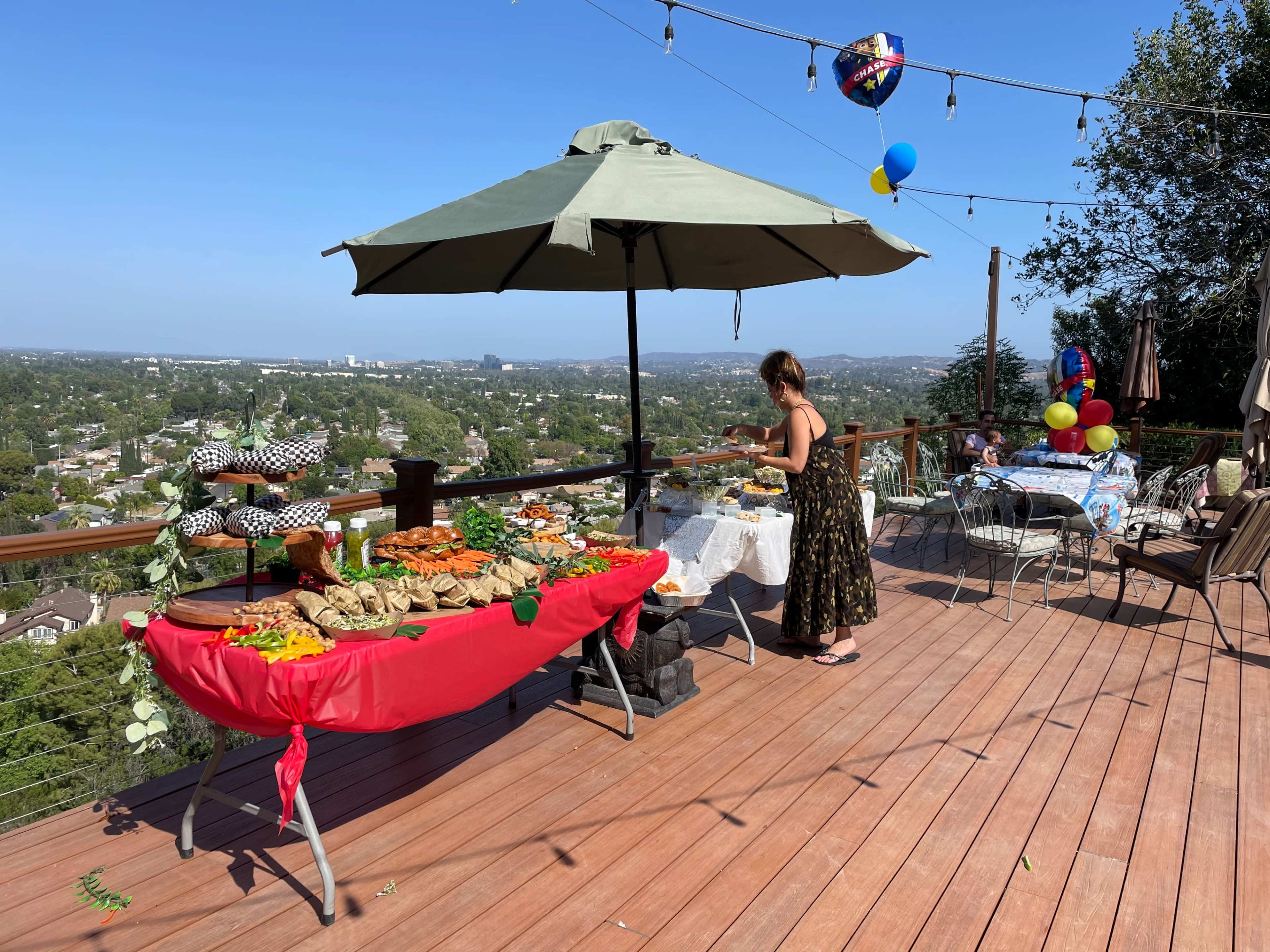 A woman arranges food on a table under an umbrella on a deck overlooking a city landscape.