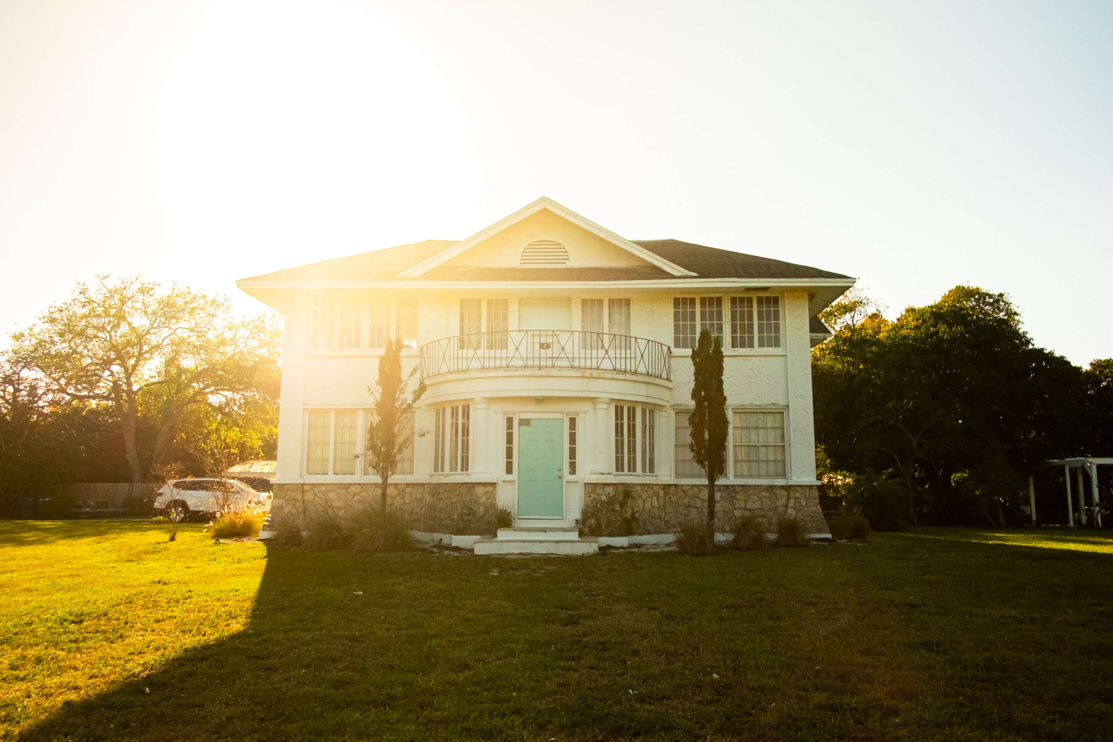 A large, two-story white house with a rounded front porch and blue door is set on a grassy lawn under bright sunlight.