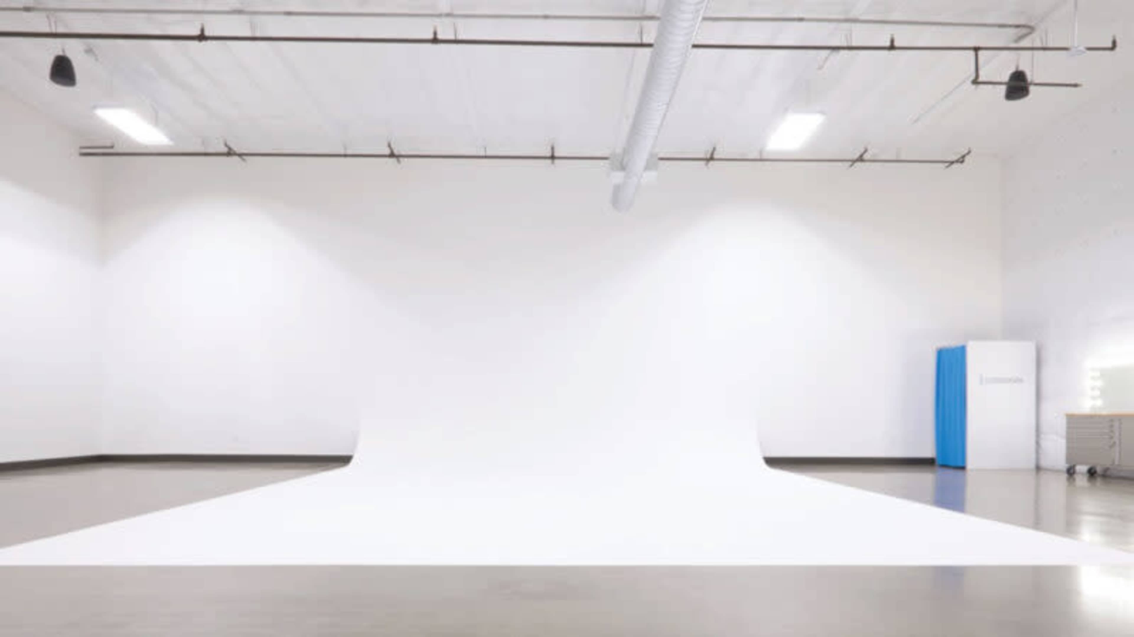 A spacious studio features a seamless white backdrop extending onto the floor, with a mirror and light setup in the corner.