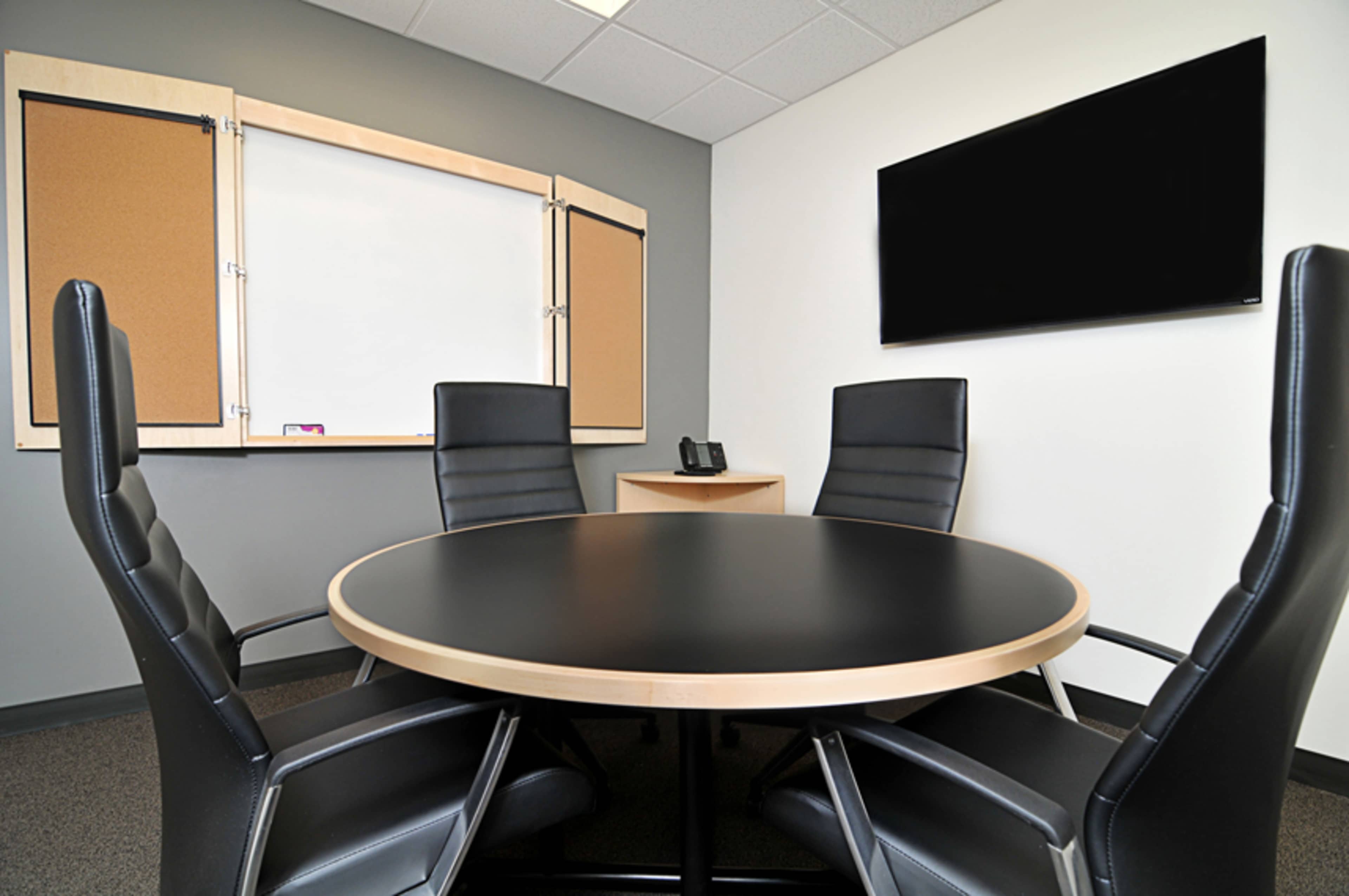 A modern conference room features a round black table surrounded by four black chairs, with a whiteboard and large screen on the walls.