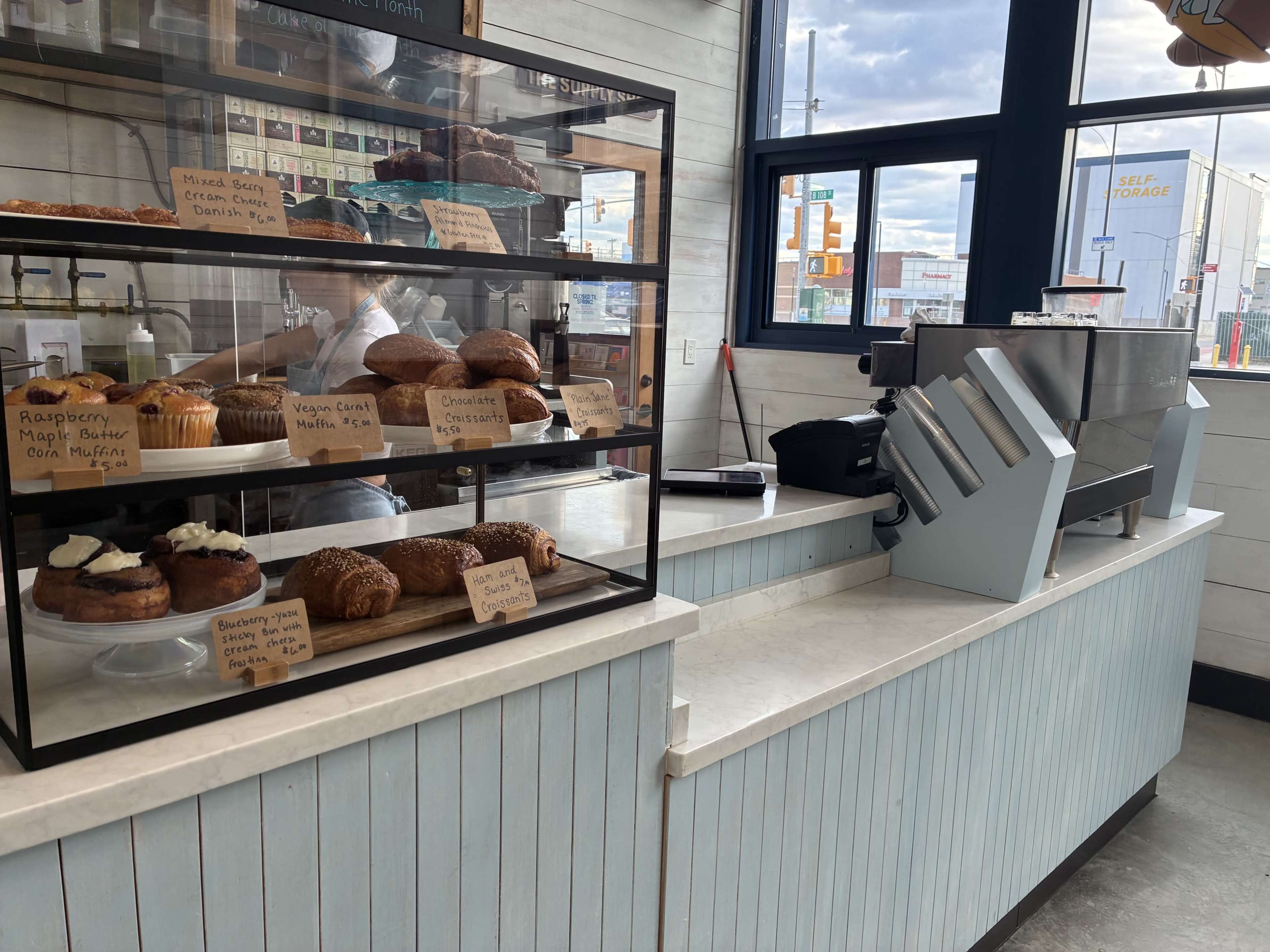 The image shows a bakery display case filled with various pastries and a coffee machine on a counter.