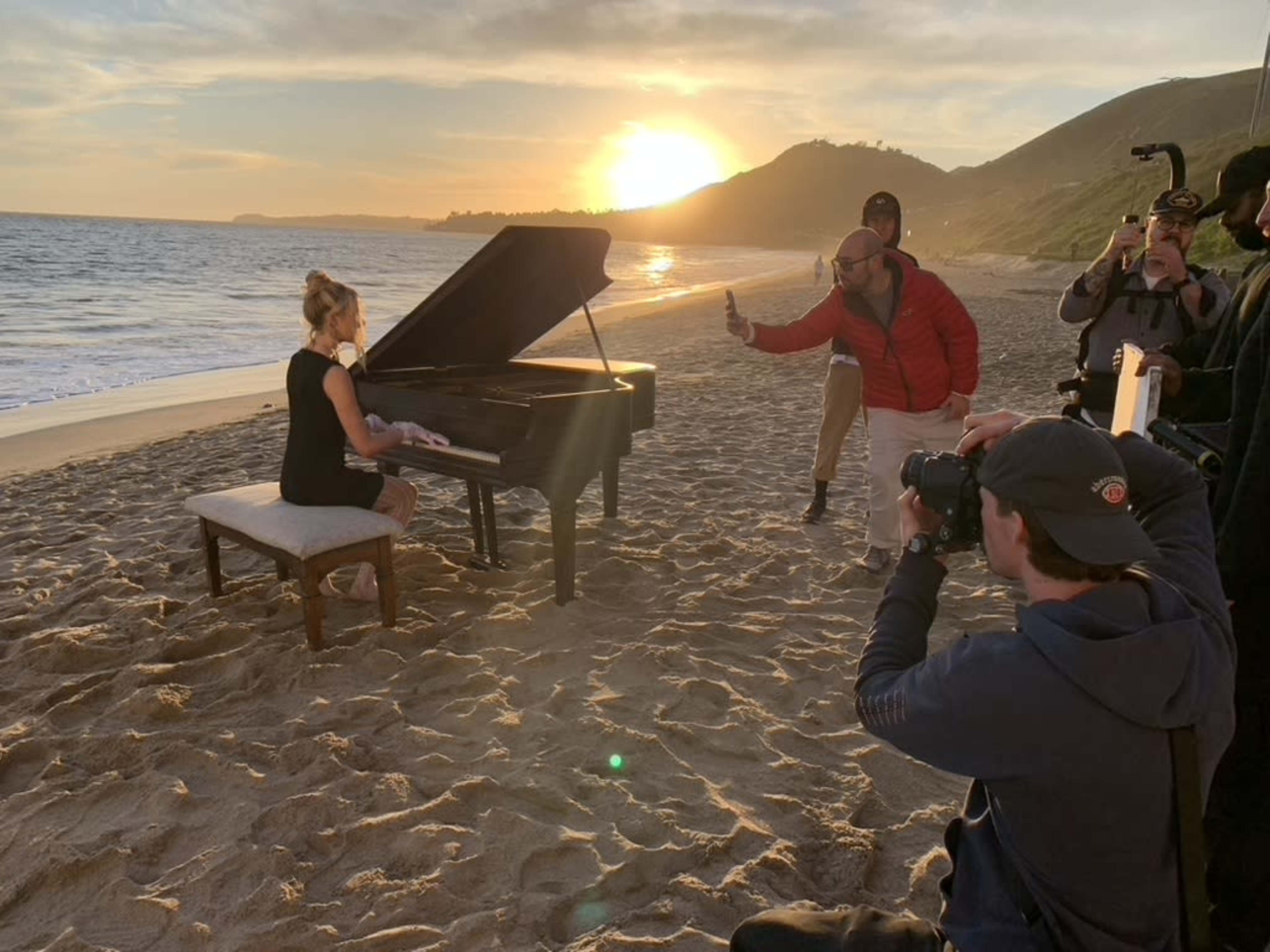 A woman plays a grand piano on the beach at sunset while a group of people observes and takes photos.