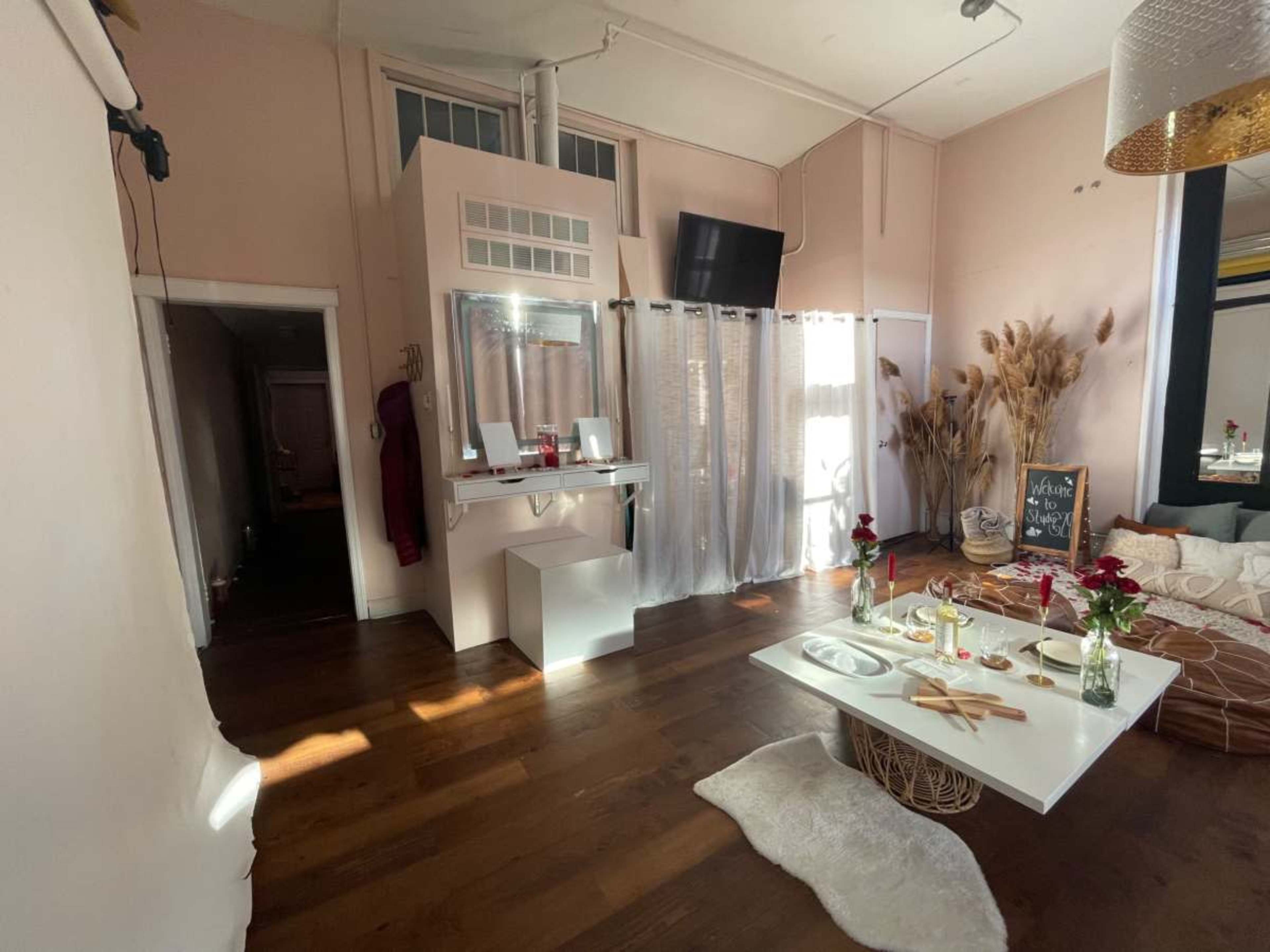 The image shows a bright, stylish interior with a white table set for dining, decorative plants, and a large mirror against a pink wall.