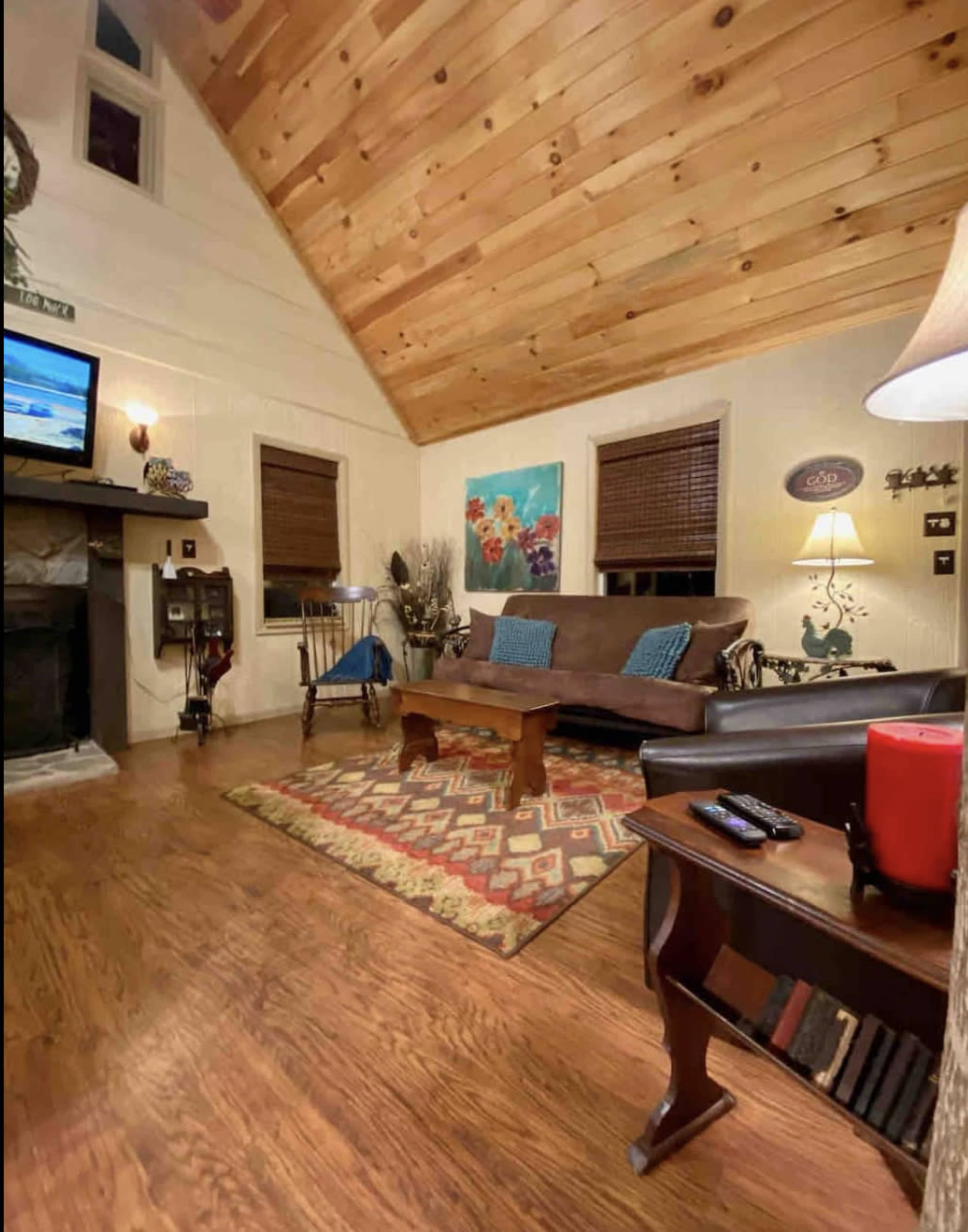 A cozy living room features wooden ceilings, a brown leather sofa, and a colorful artwork on the wall, with a coffee table and a patterned rug on the wooden floor.