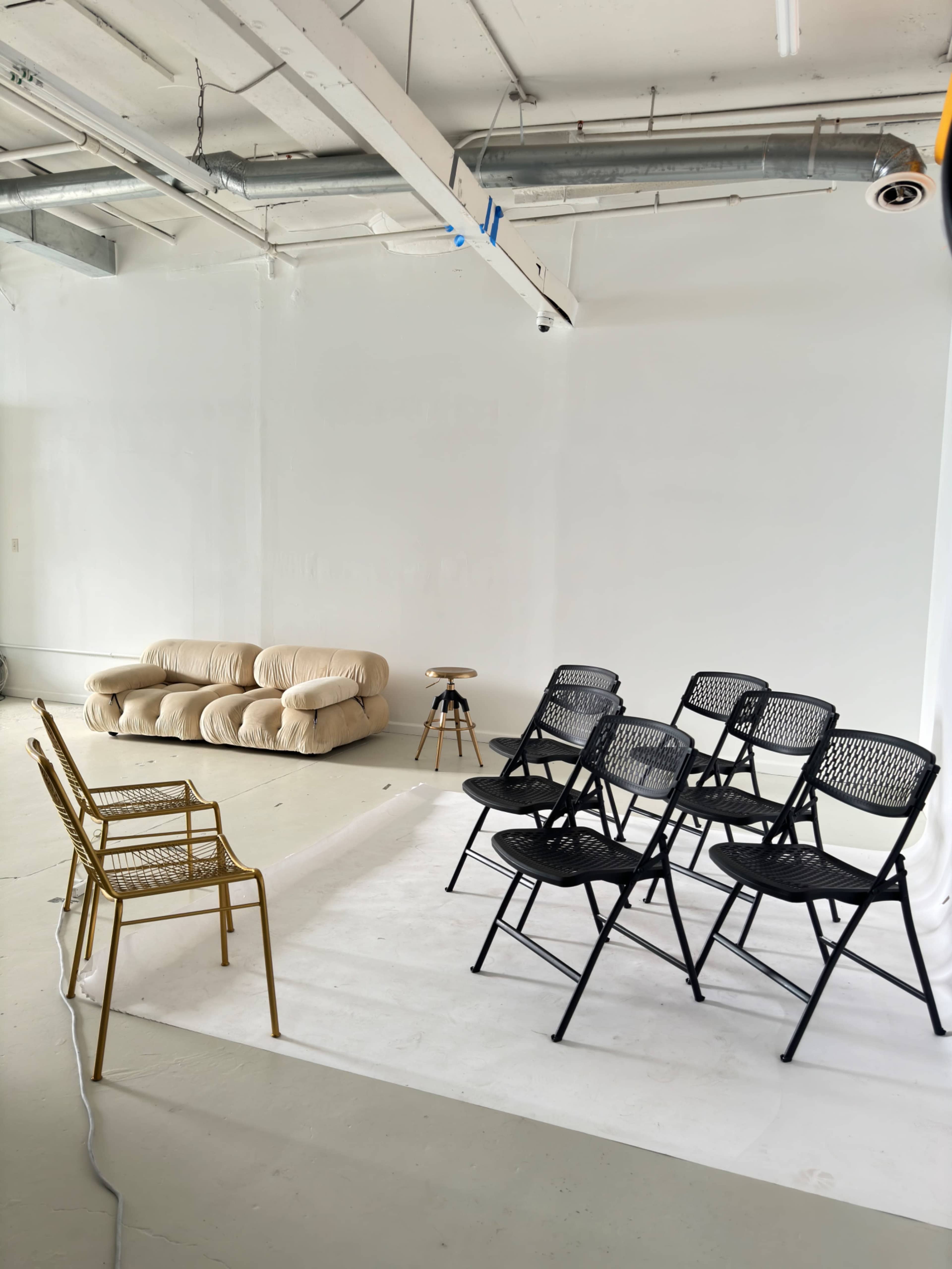 A minimalist room with a beige couch, a beige stool, and a row of black folding chairs arranged on a white backdrop.