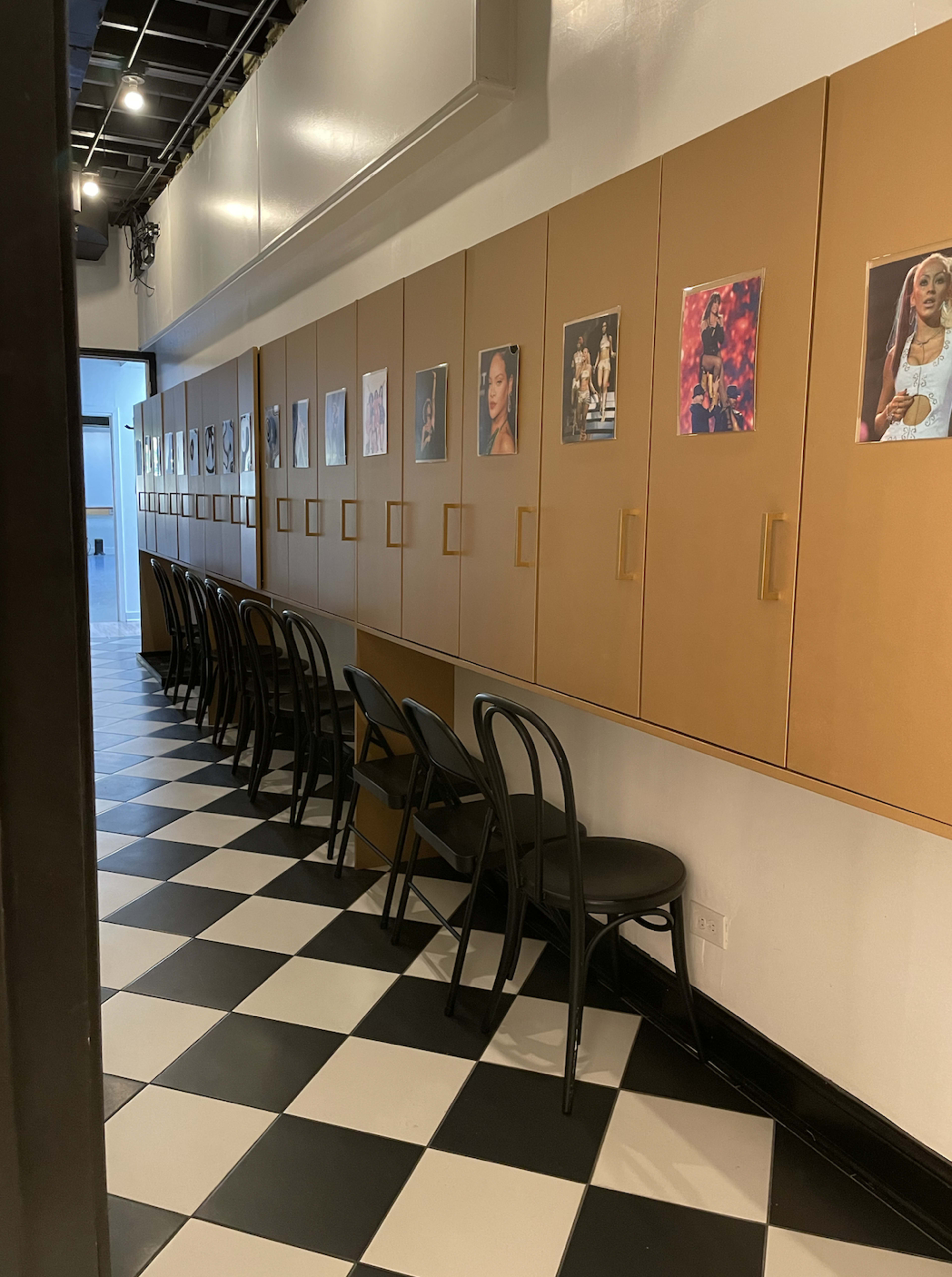 A long corridor features black and white checkered flooring with rows of chairs facing a wall of cabinets adorned with framed photographs.