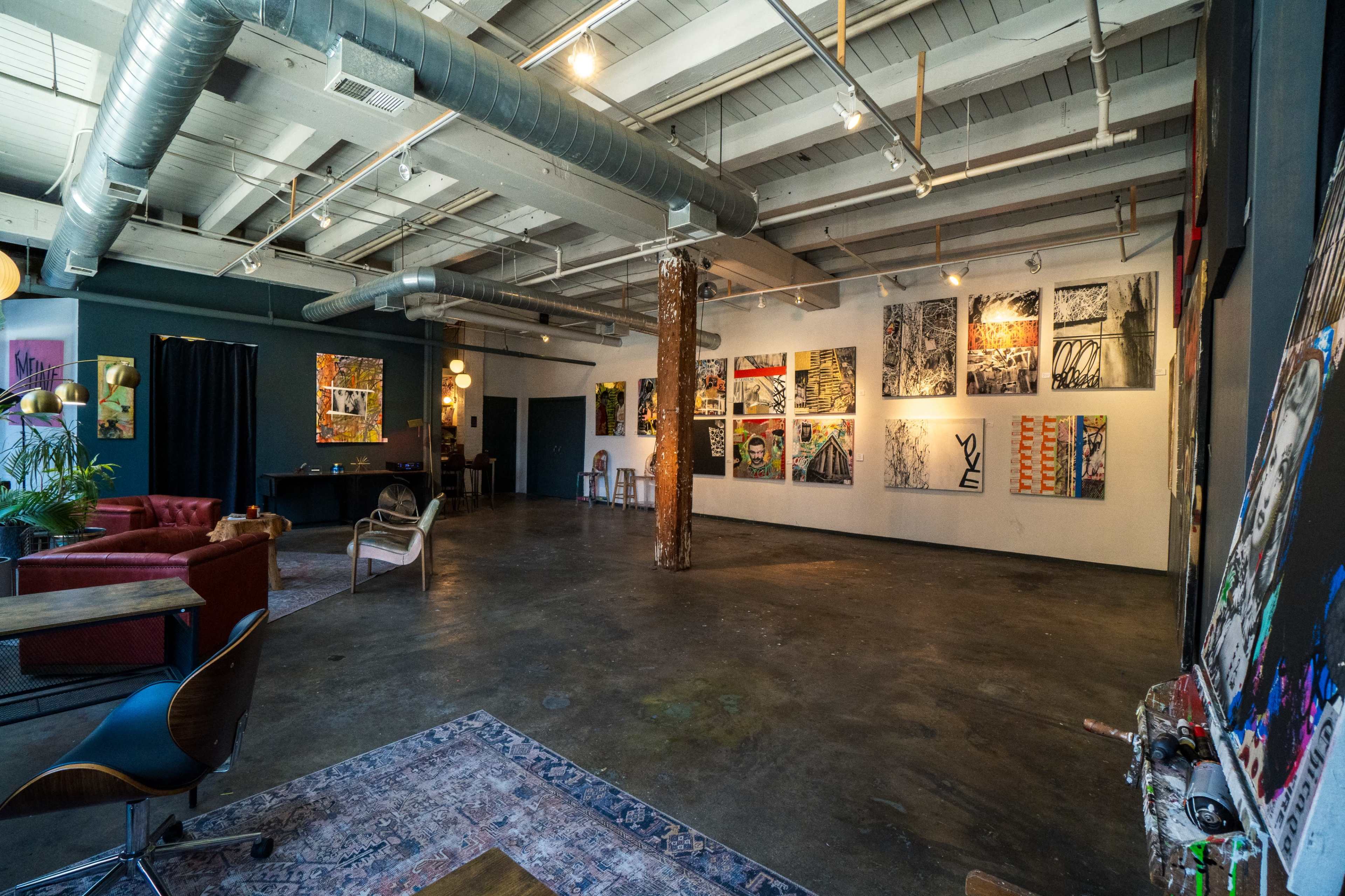 An art gallery featuring several colorful paintings on the walls, with a seating area and exposed industrial elements.