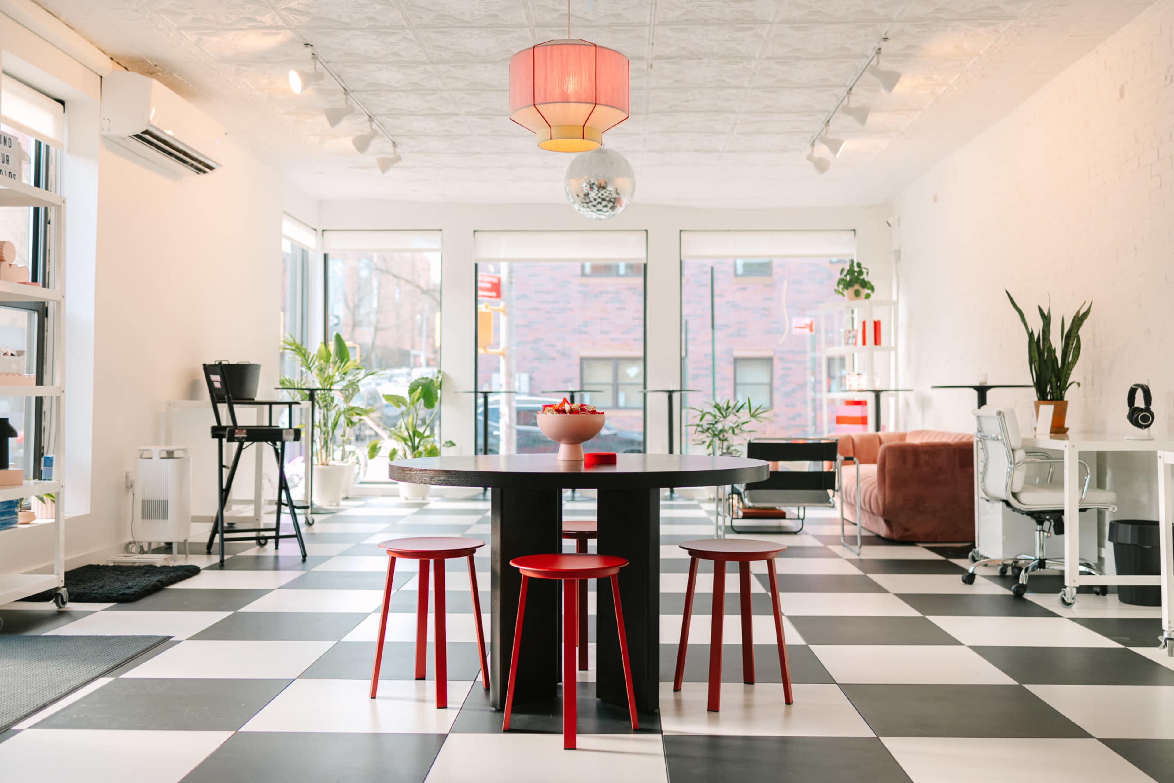 A modern room features a black and white checkered floor, a round table with three red chairs, and large windows filled with plants and natural light.