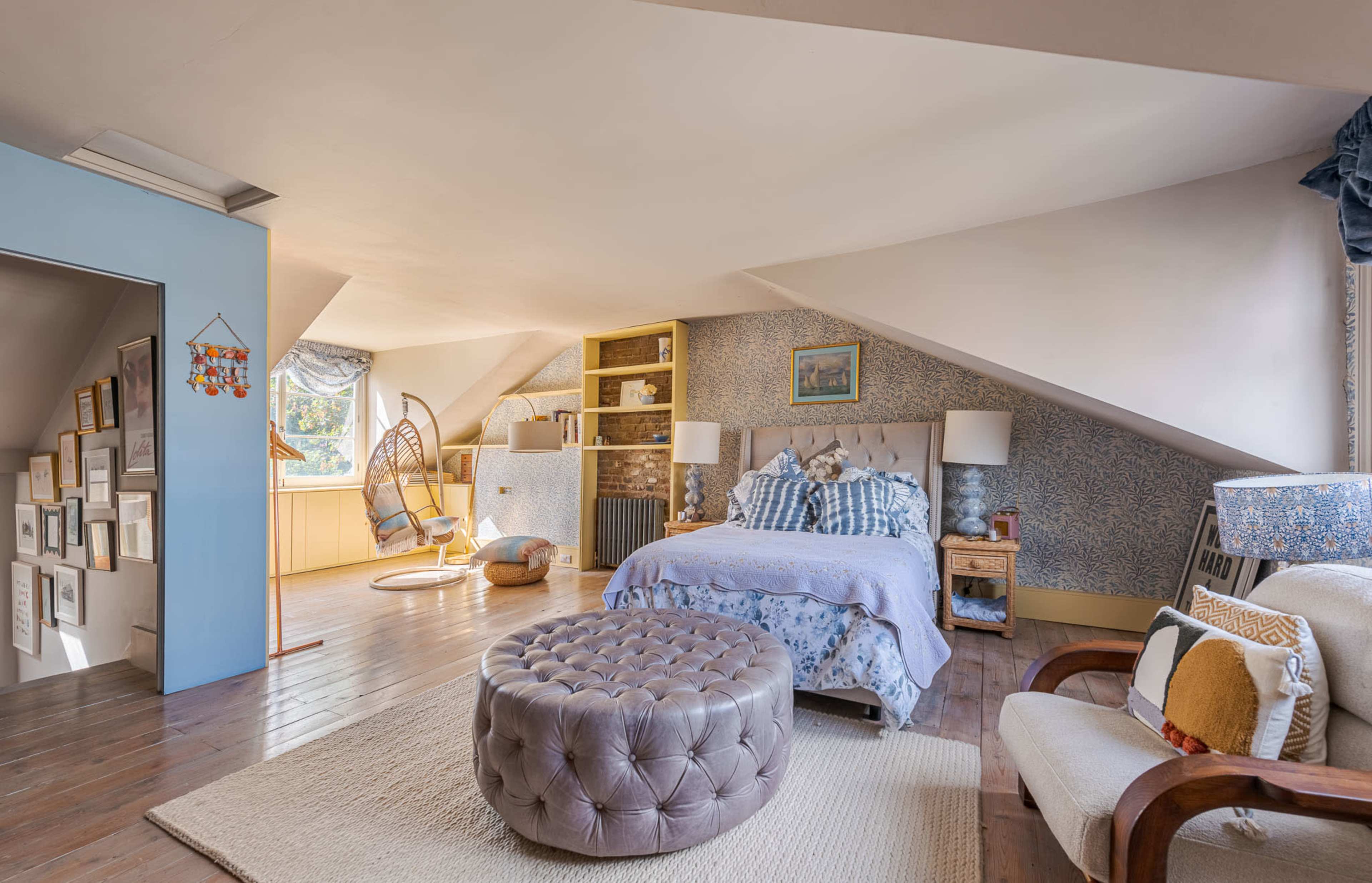 The image shows a cozy attic bedroom with sloped ceilings, a bed, a round ottoman, and a nook featuring a hanging chair.