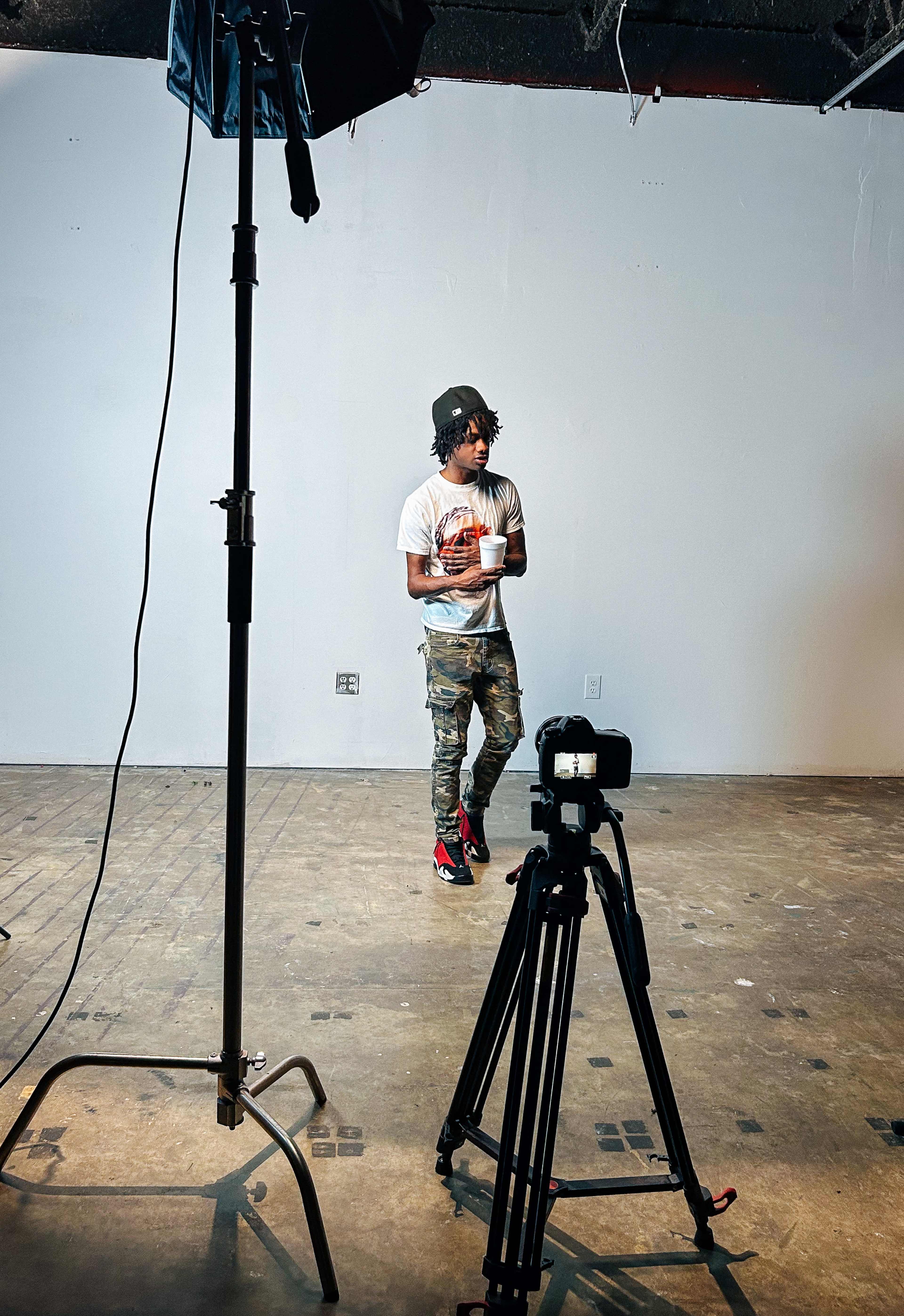 A person stands in a sparse room with a camera and lighting setup, wearing casual clothing and holding a drink.